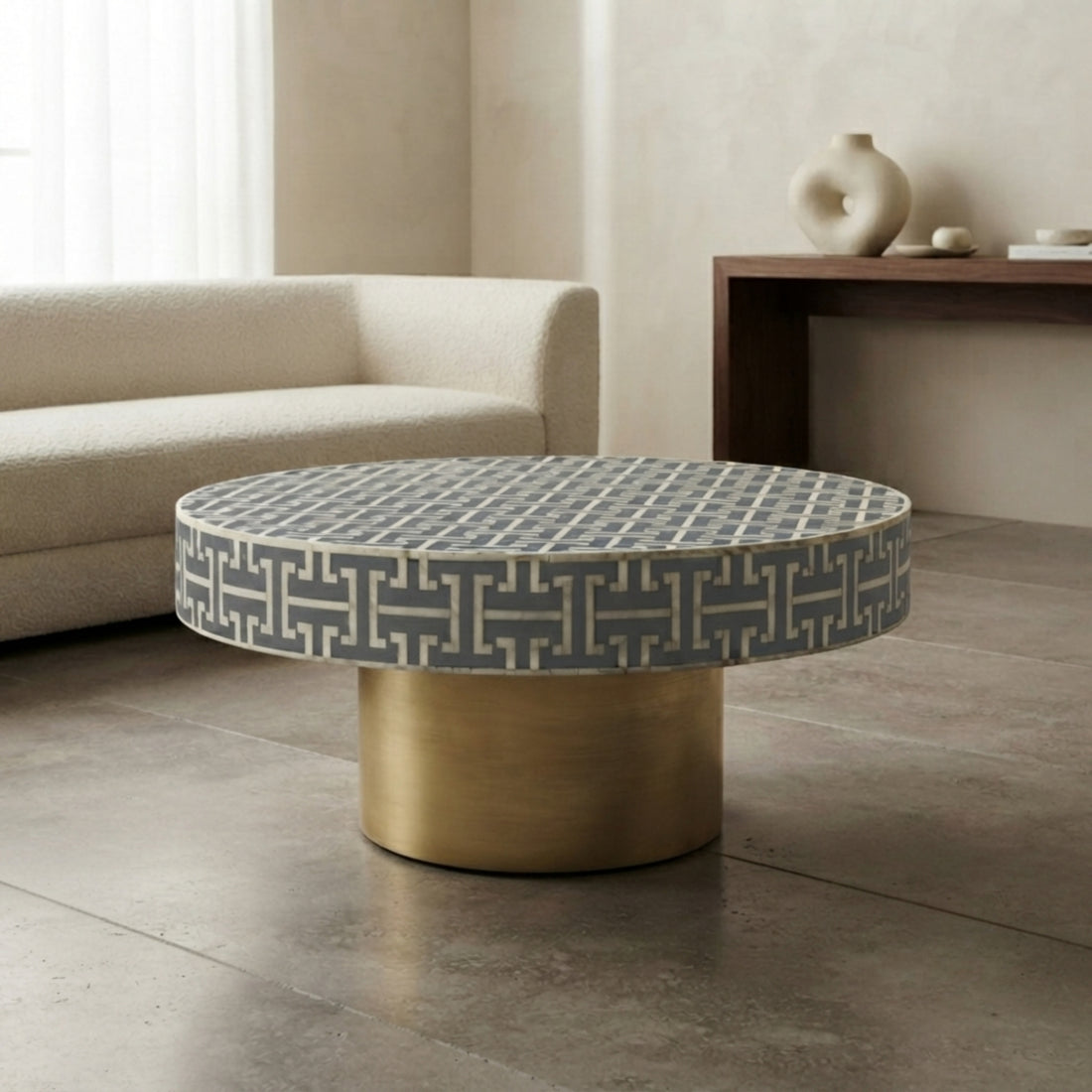 A round coffee table with a gray and white geometric patterned top and a cylindrical brushed gold base, styled in a modern living room setting.