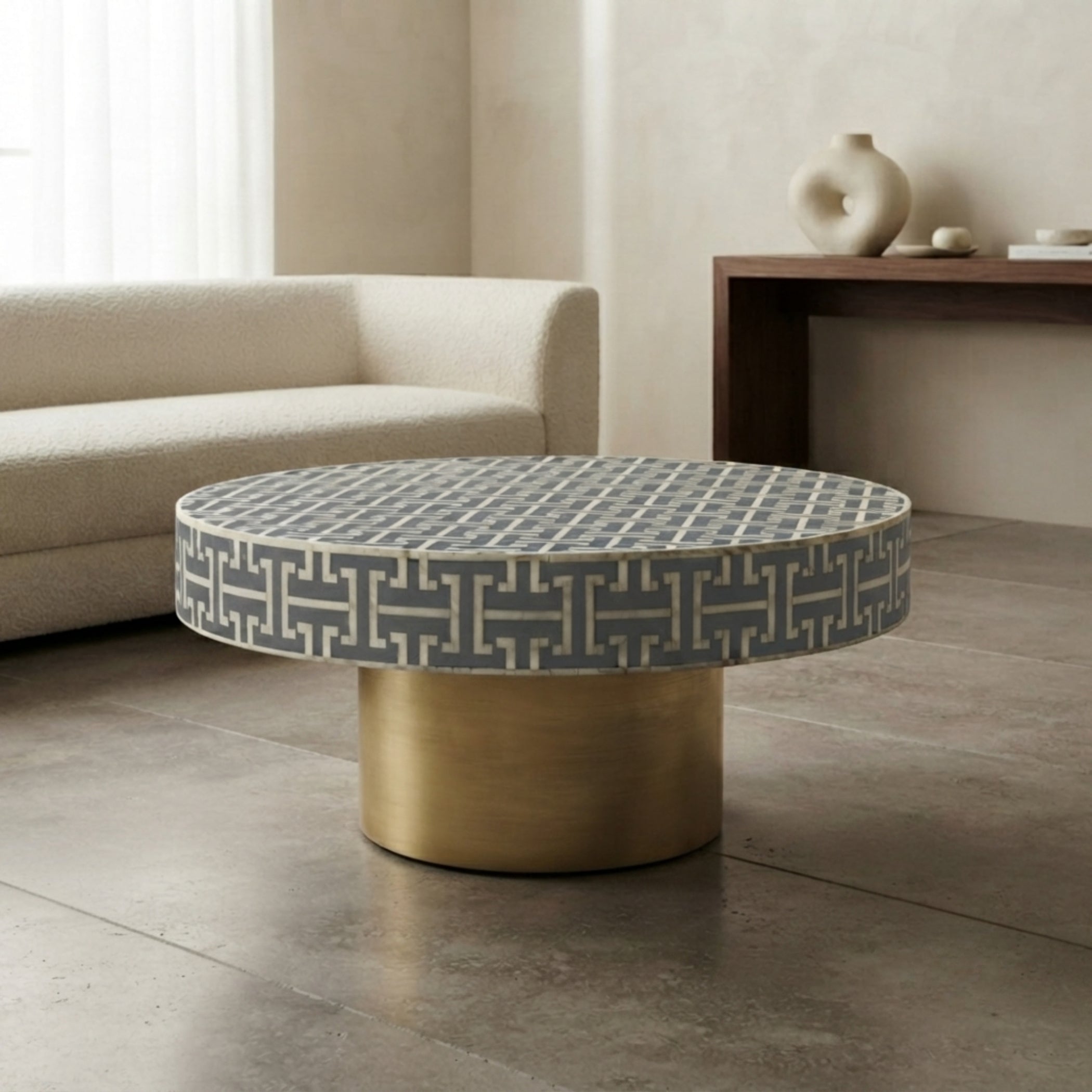 A round coffee table with a gray and white geometric patterned top and a cylindrical brushed gold base, styled in a modern living room setting.
