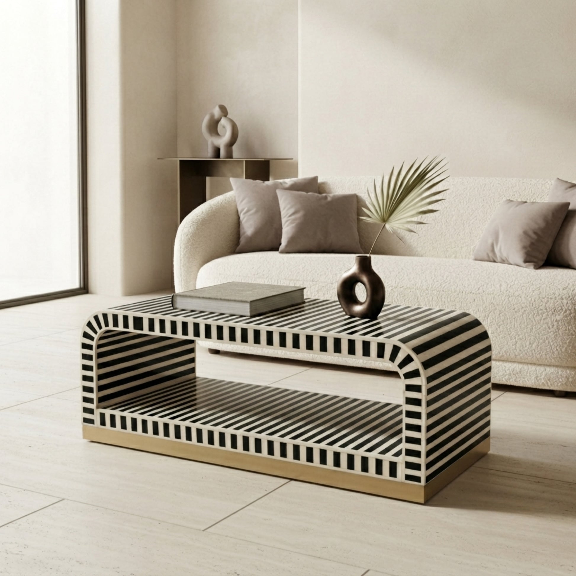 A modern rectangular coffee table with a black and white striped bone-and-wood frame and a gold metal base, styled in a minimalist living room next to a cream bouclé sofa.