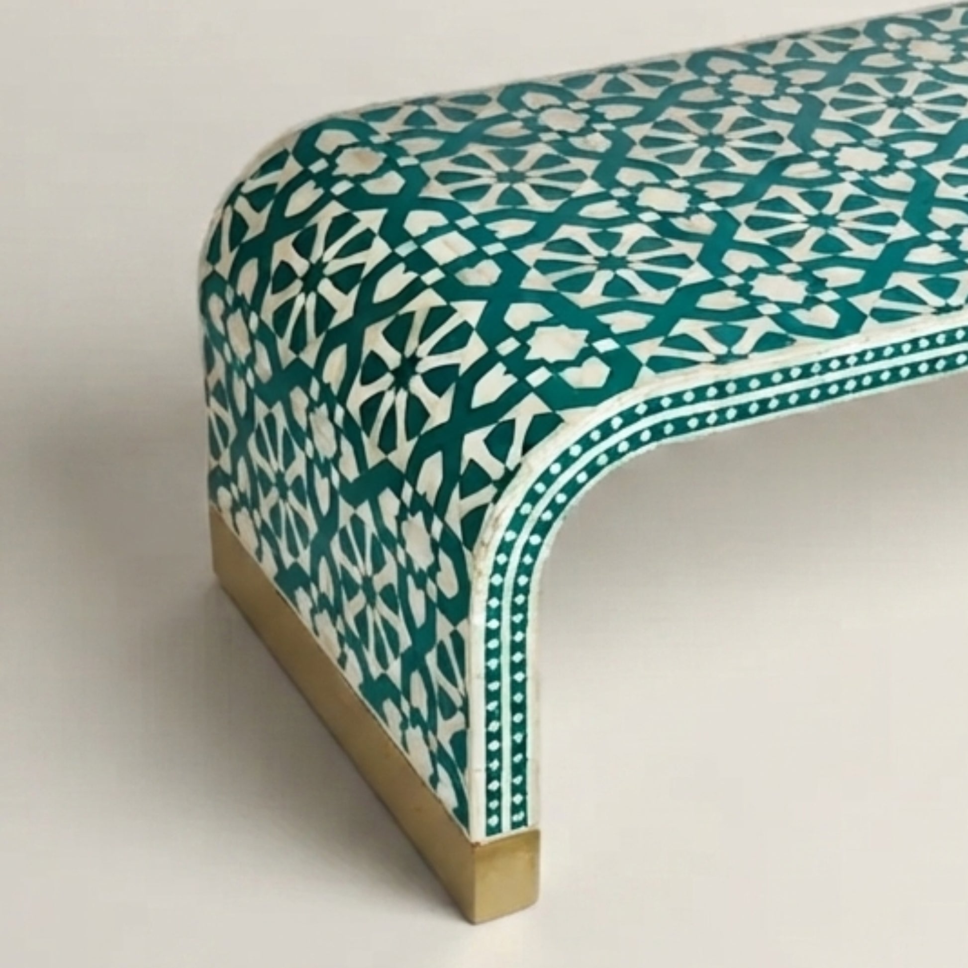 A close-up of a modern curved bench featuring a teal and white geometric inlay pattern with a brass-colored metal base.