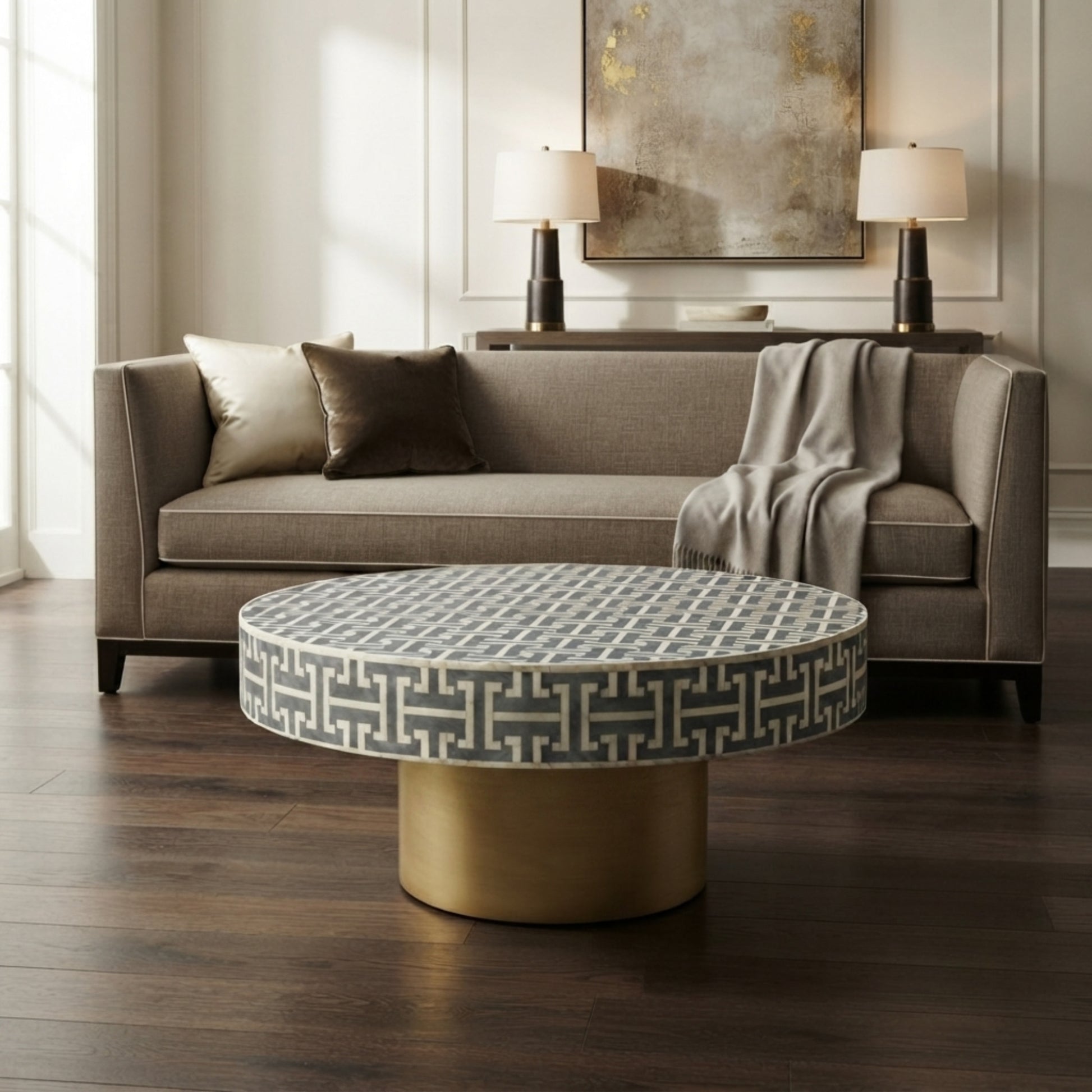 A round coffee table with a gray and white geometric patterned top and a cylindrical gold base, positioned in front of a gray fabric sofa in a modern living room setting.