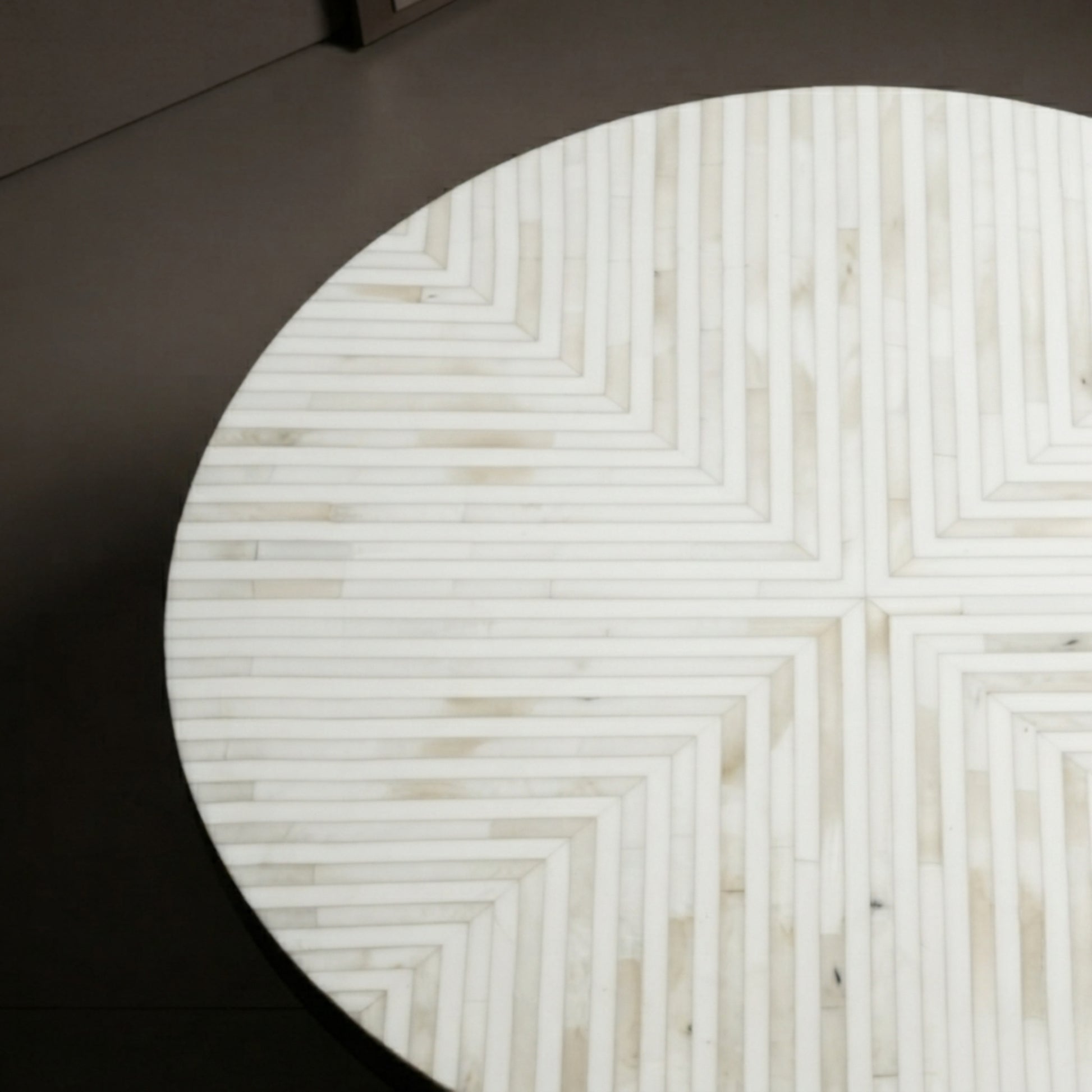 A round dining table with a white and beige chevron inlay pattern, shown from a top-down angle against a dark gray floor.