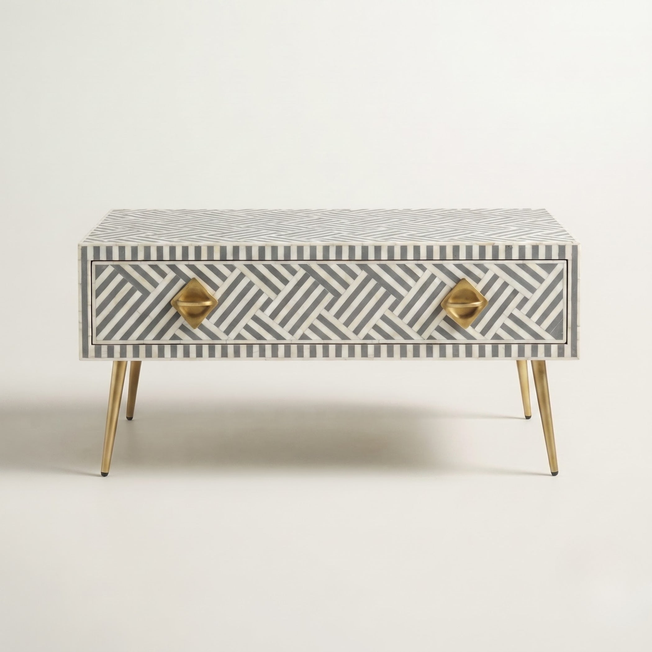 A mid-century modern single-drawer side table featuring a gray and white geometric bone-inlay pattern, gold geometric drawer pulls, and tapered gold metal legs, set against a plain off-white background.