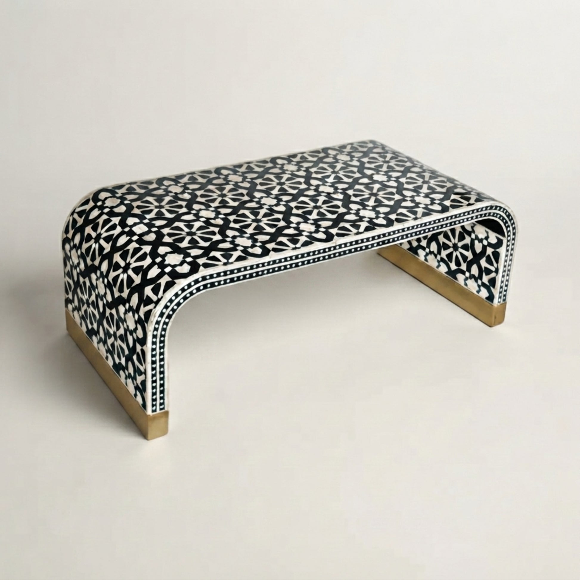 A curved bench with a black and white geometric bone-inlay pattern and gold-toned metal legs, photographed on a clean off-white background.