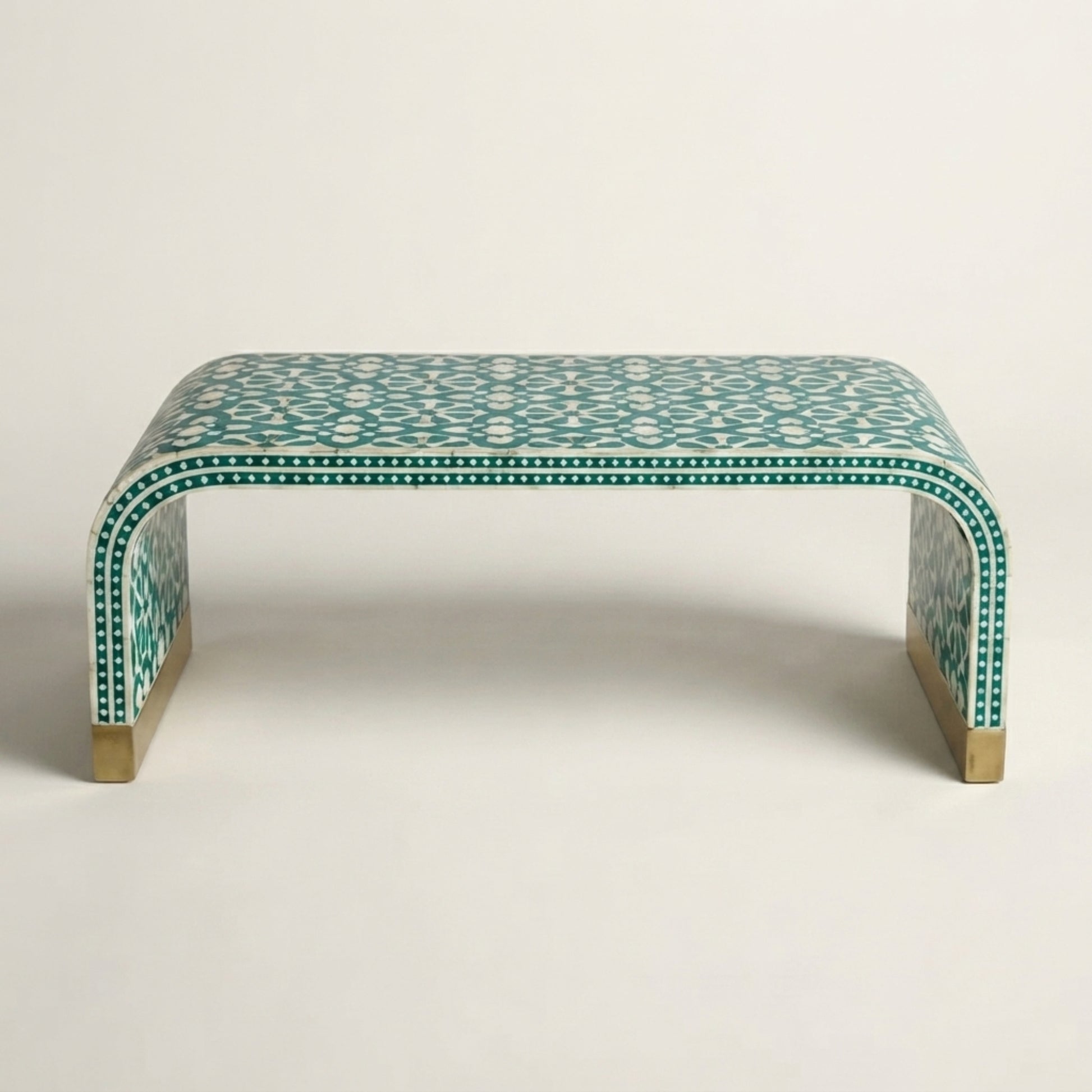 A curved bench with a green and white geometric bone-inlay pattern and brass-colored legs, photographed against a neutral light gray background.