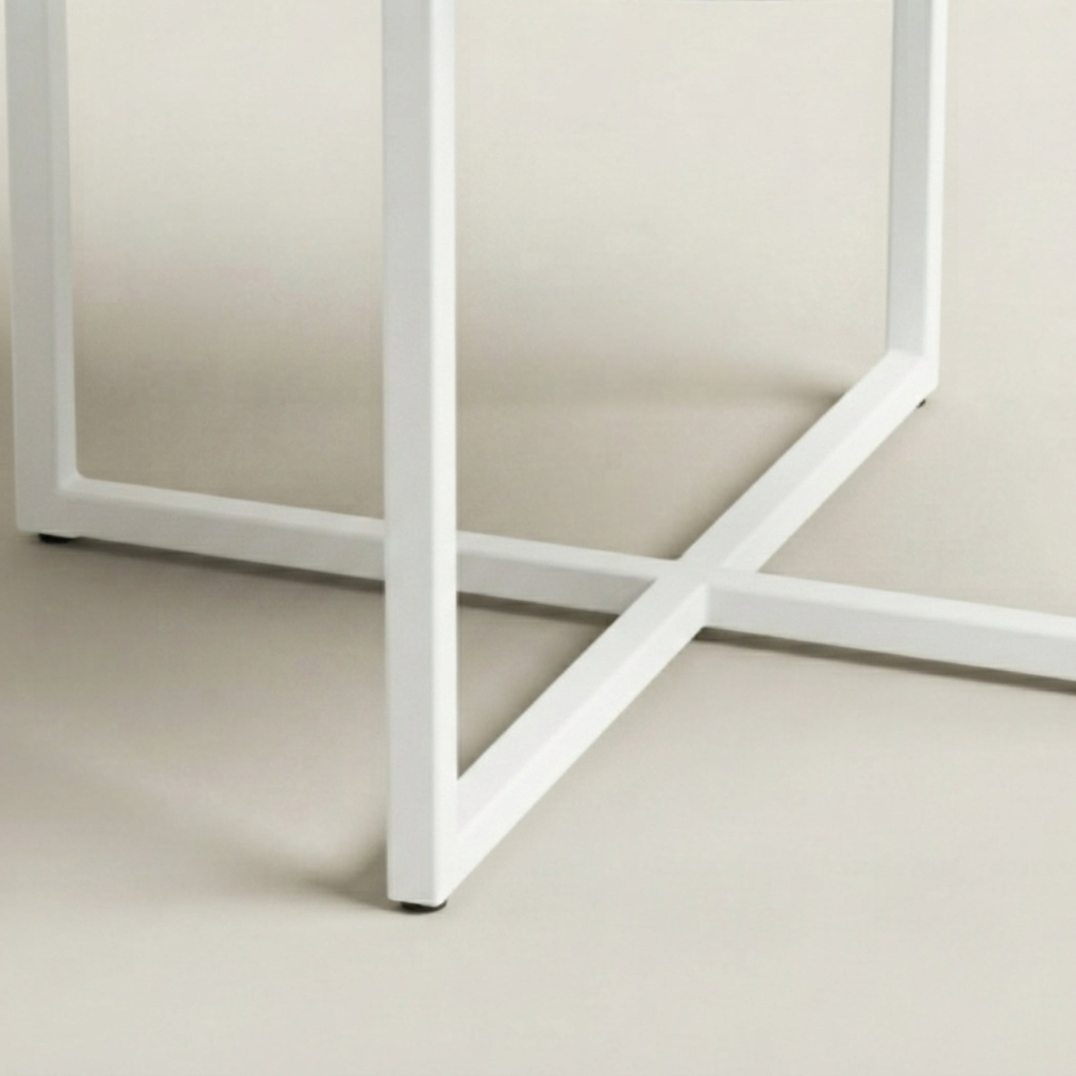 Close-up of a white metal table base with a square frame and intersecting crossbar, shown on a light beige background.