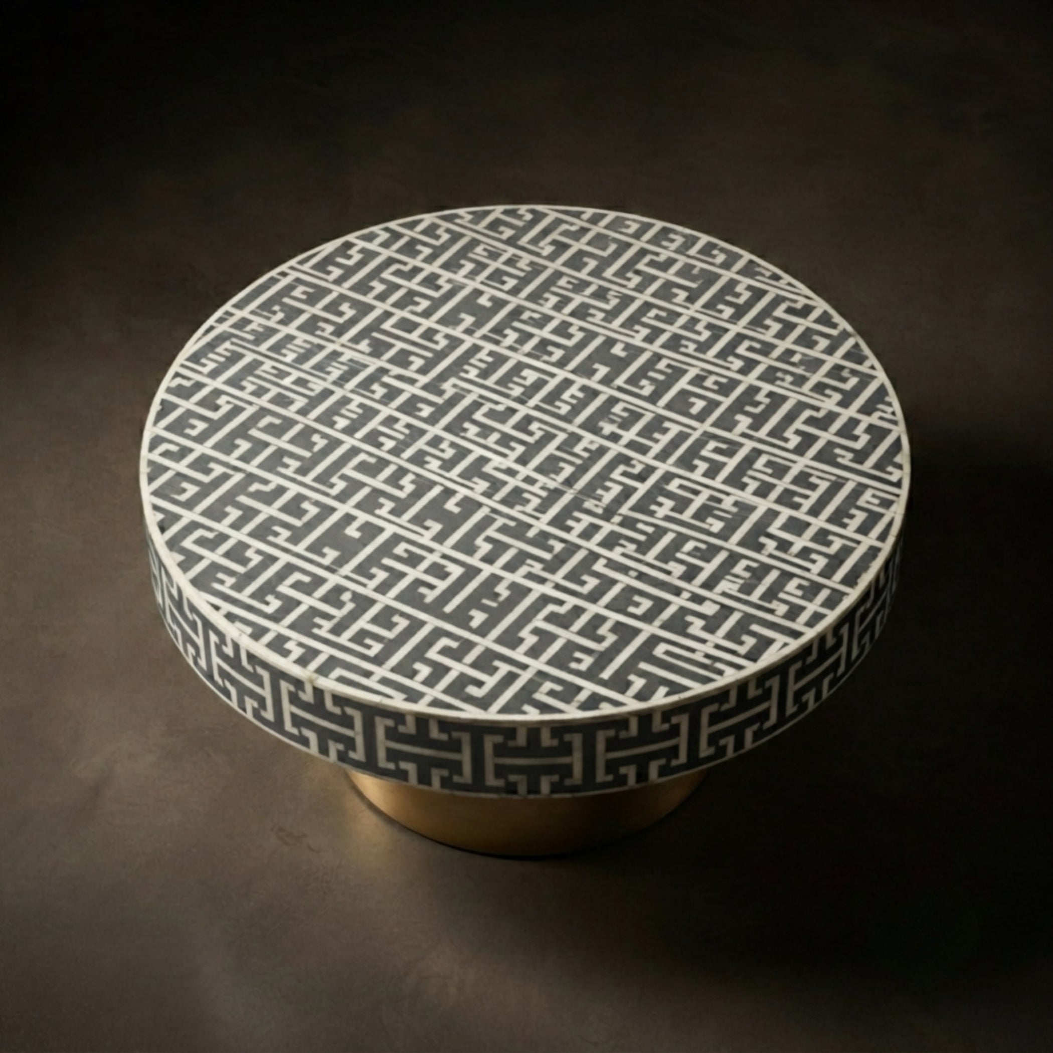 A round side table with a white top featuring a dark gray geometric Greek key pattern, supported by a round gold-colored base, set against a dark brown background.