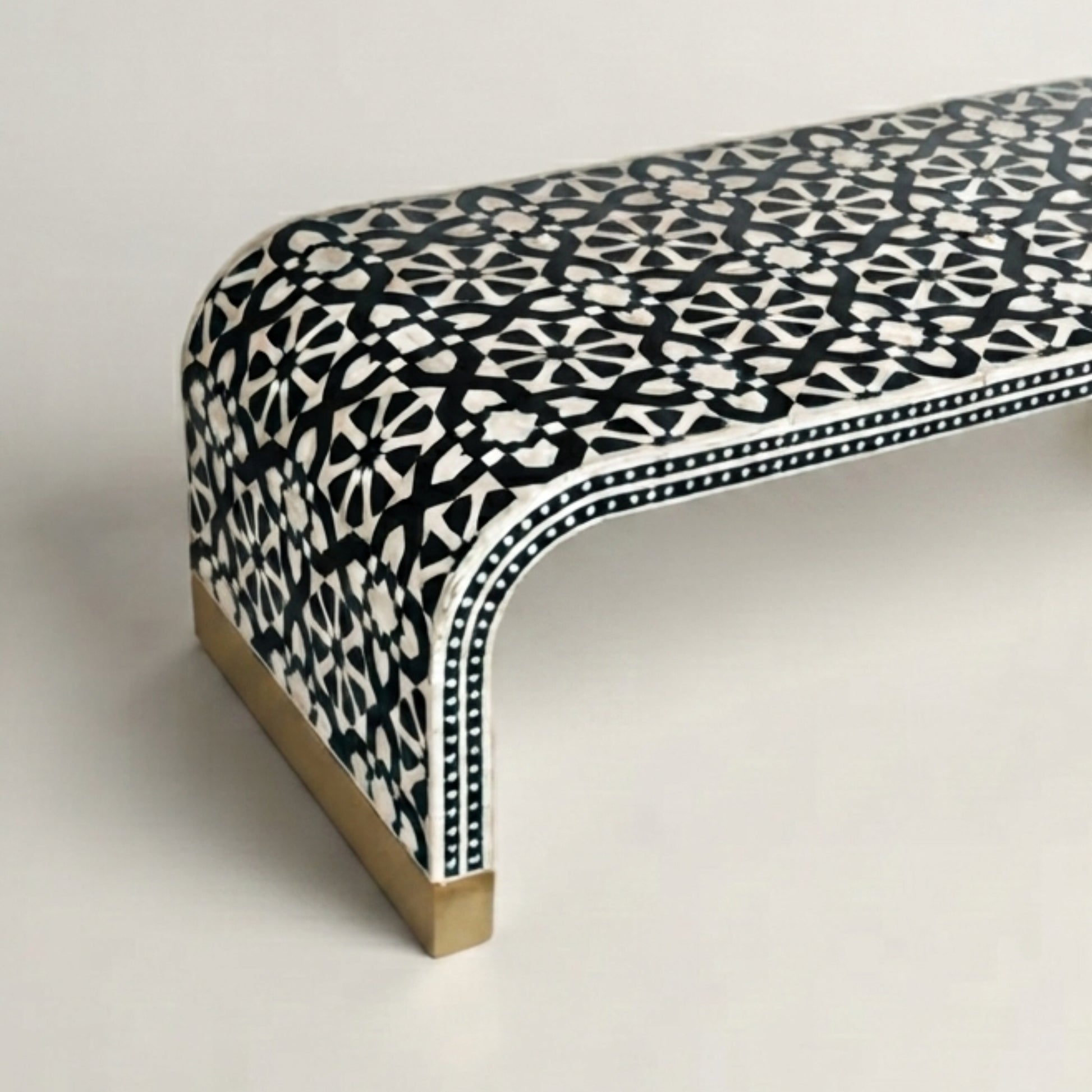 A close-up of a curved bench upholstered in a black and white geometric patterned fabric, with a light wood base and brass nailhead trim, set against a plain light gray background.