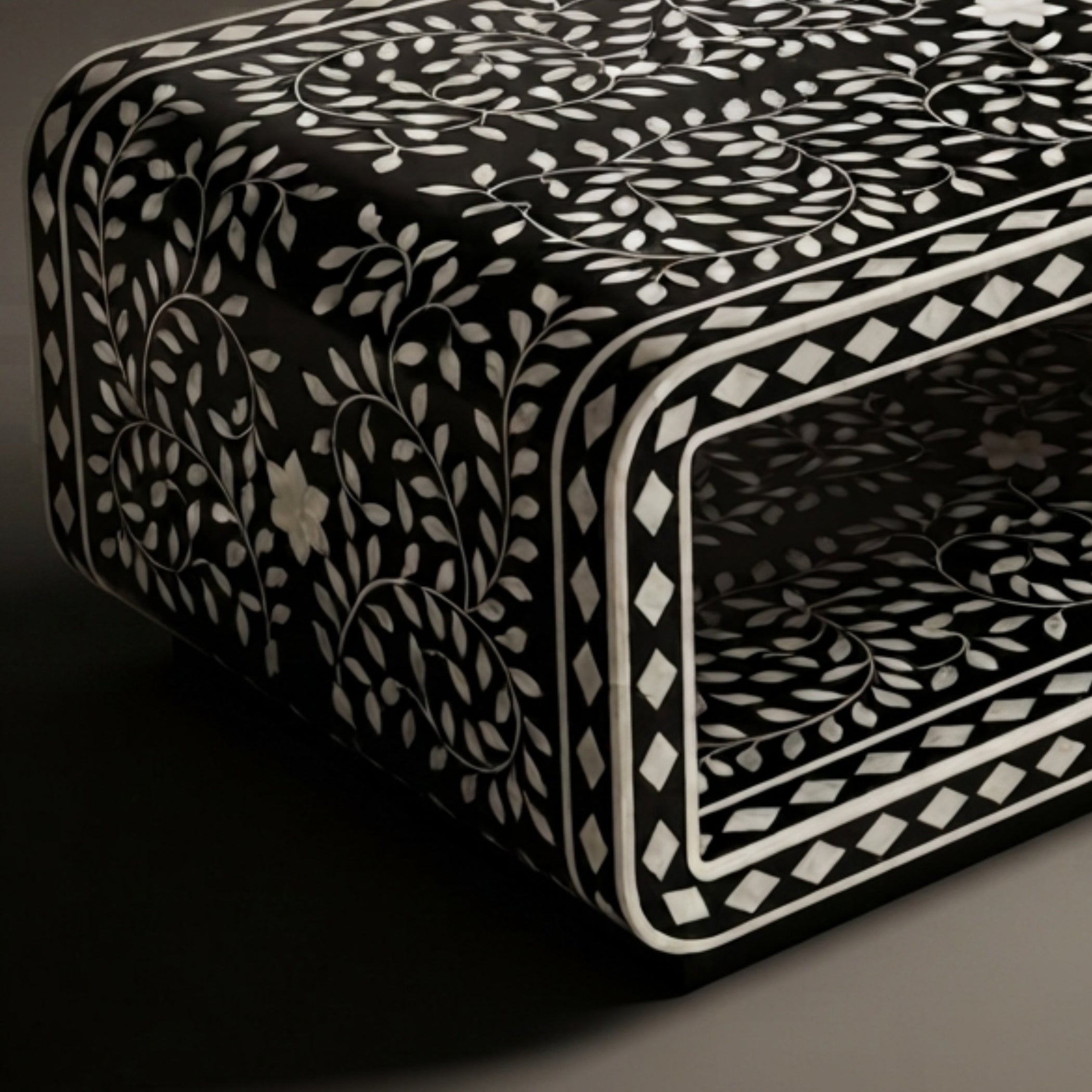 A close-up of a black rectangular box or furniture piece decorated with an intricate white bone-inlay pattern of leaves, vines, and geometric borders.