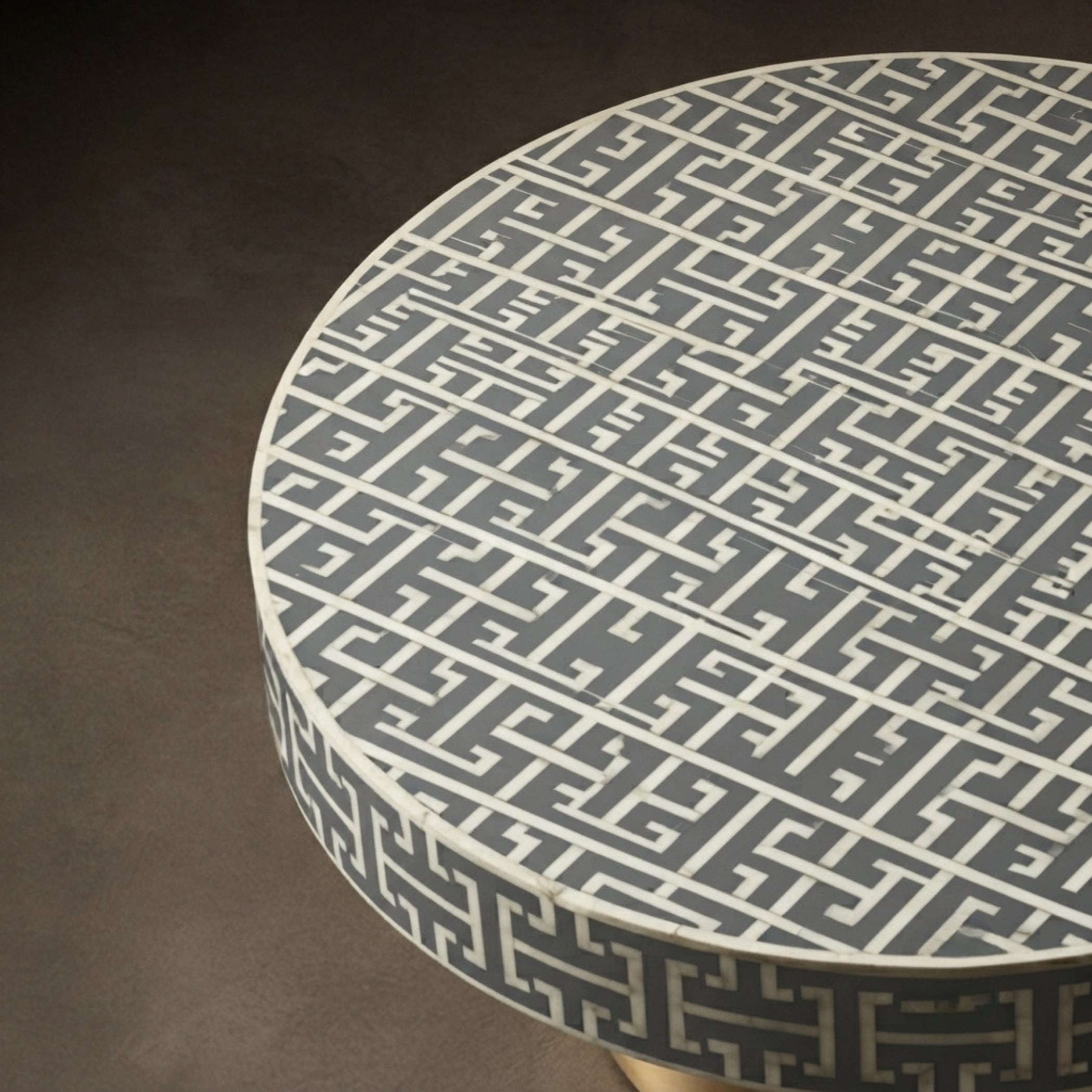 A close-up of a round table with a dark gray and white geometric maze pattern on the tabletop and side, set against a dark brown background.