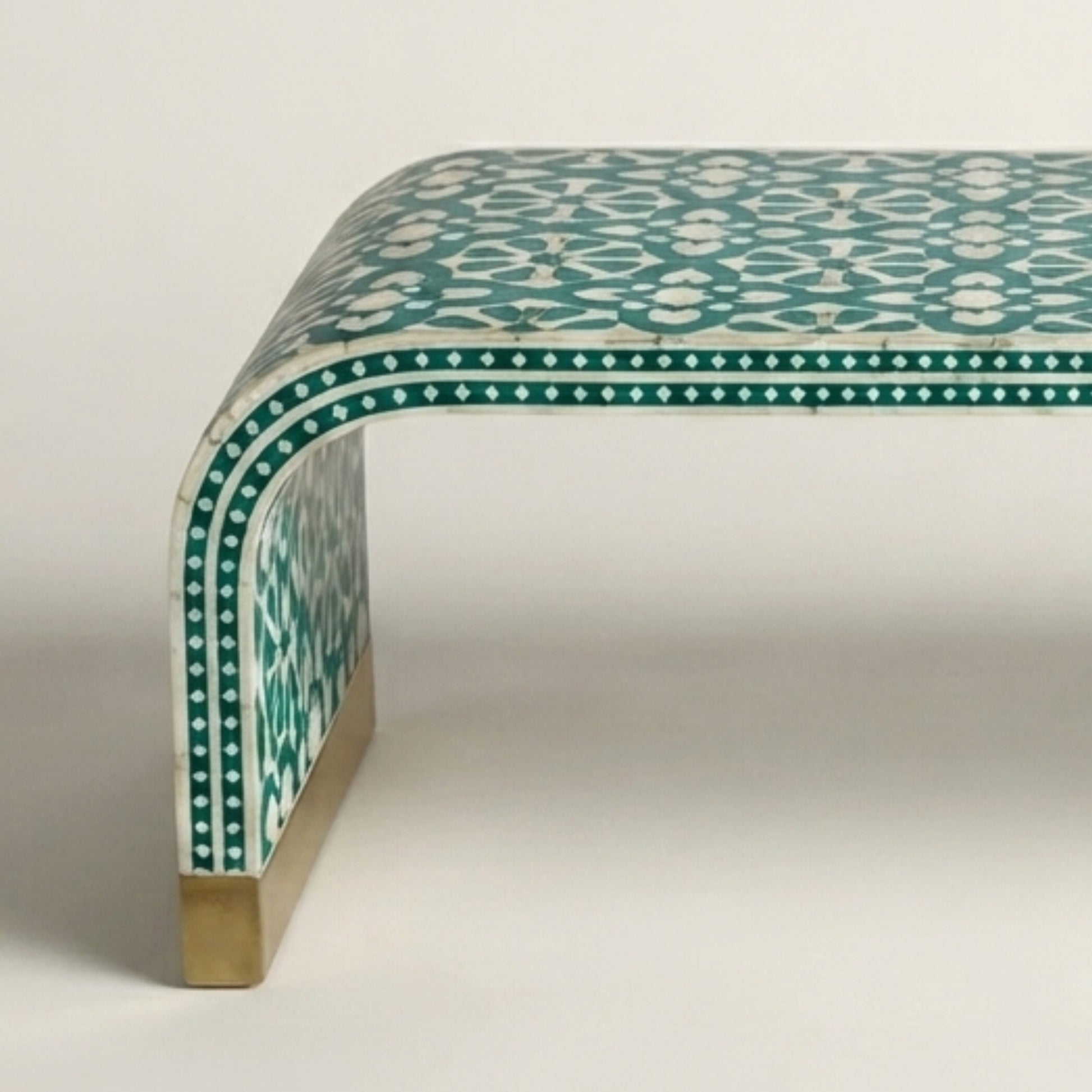 A close-up of a modern bench with a curved backrest, featuring a teal and white geometric patterned inlay and a brass-colored metal leg.