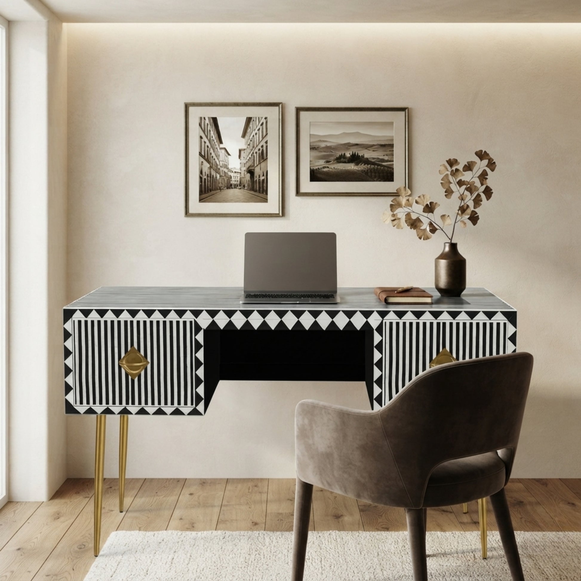 A modern black writing desk with geometric black and white striped drawer fronts and gold legs, styled in a home office setting with a gray velvet chair, laptop, and framed wall art.