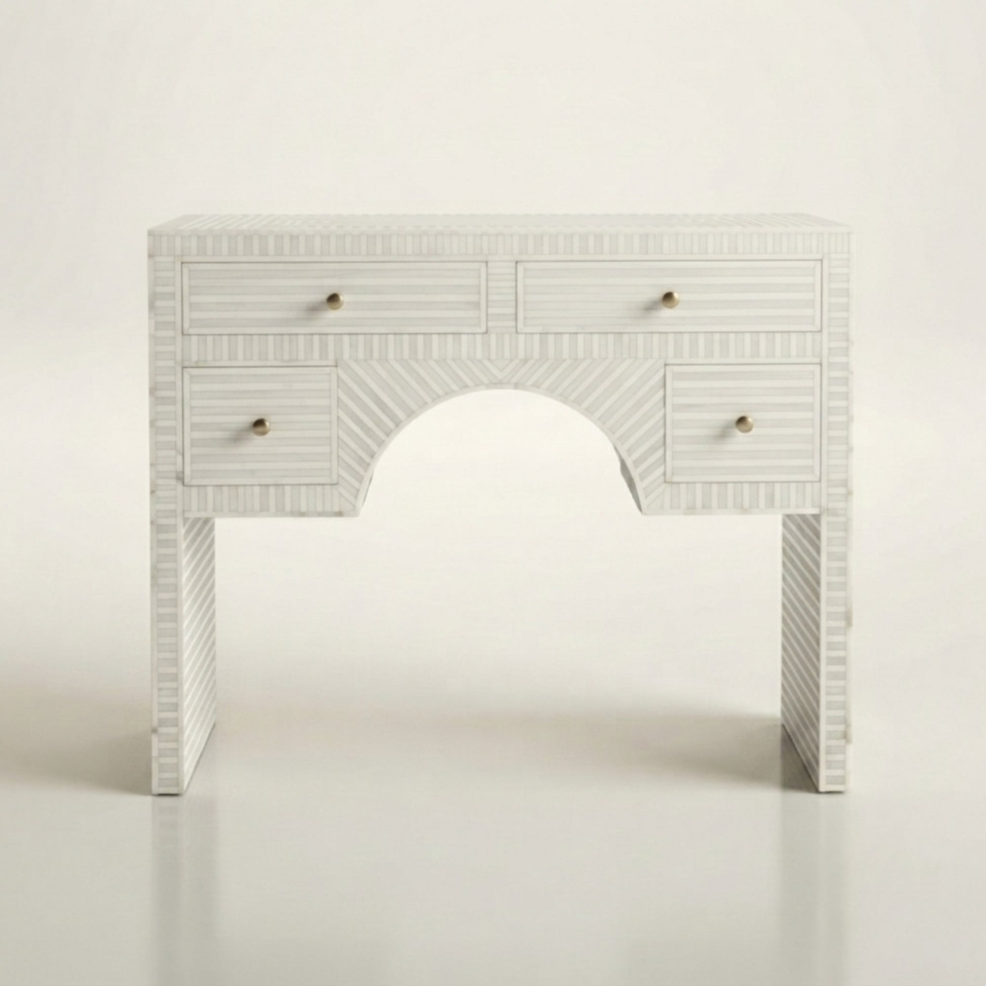 A white carved console table with a central arched opening, featuring four drawers with small brass knobs and a textured chevron pattern on the legs, set against a plain white background.