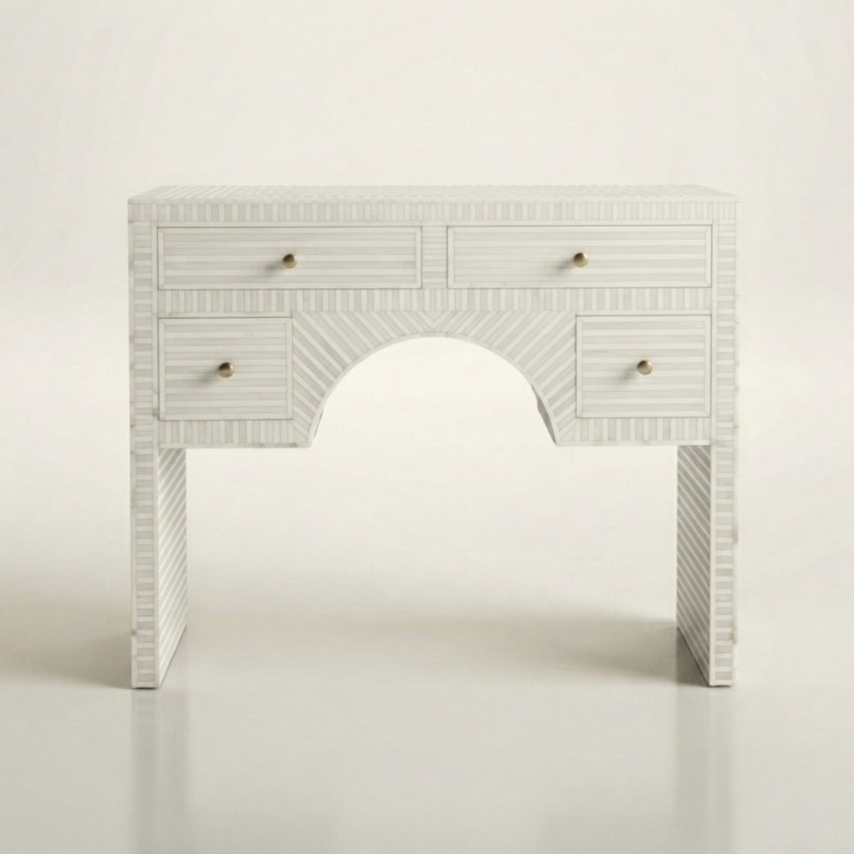 A white carved console table with a central arched opening, featuring four drawers with small brass knobs and a textured chevron pattern on the legs, set against a plain white background.