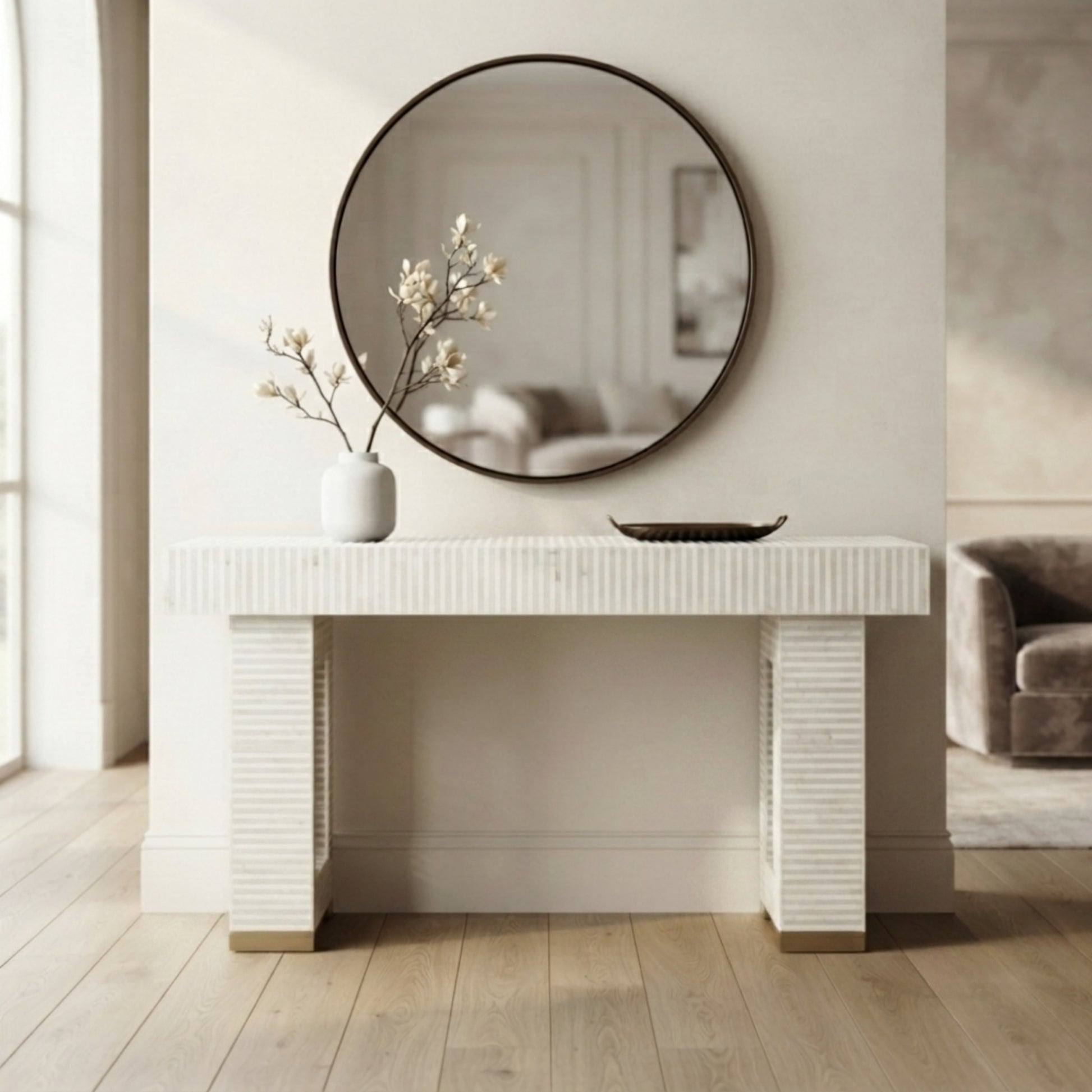 A white console table with fluted legs and a matching round wall mirror, styled in a bright, modern entryway with a small vase of branches and a shallow bowl on top.