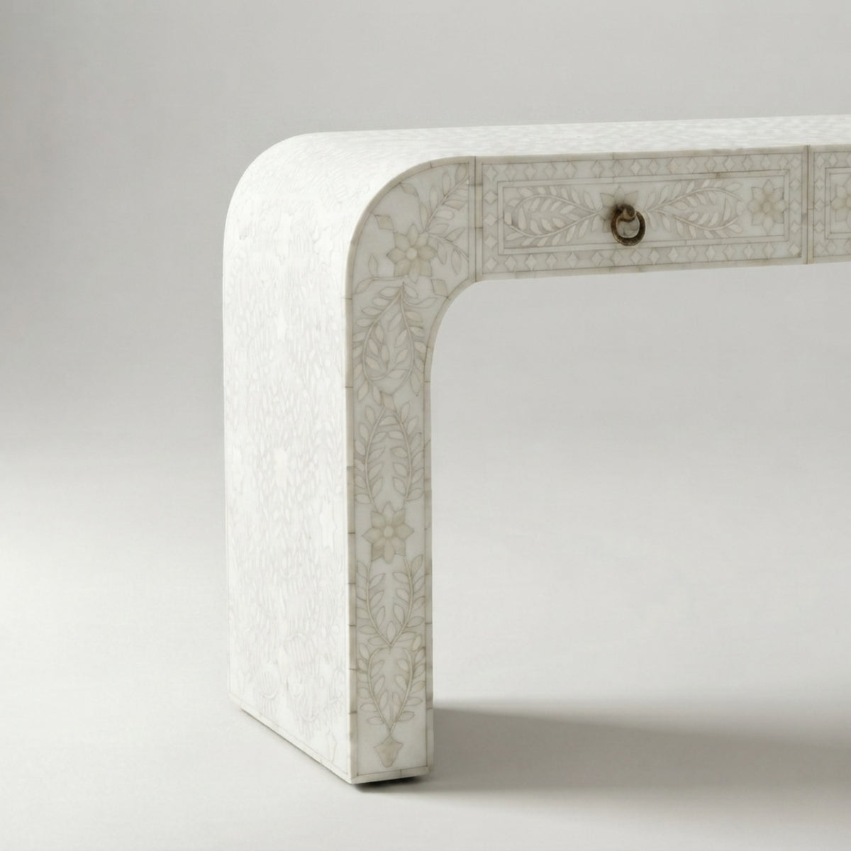 A close-up studio shot of a white table leg with delicate beige floral bone-inlay detailing and a small brass pull handle, set against a neutral light gray background.