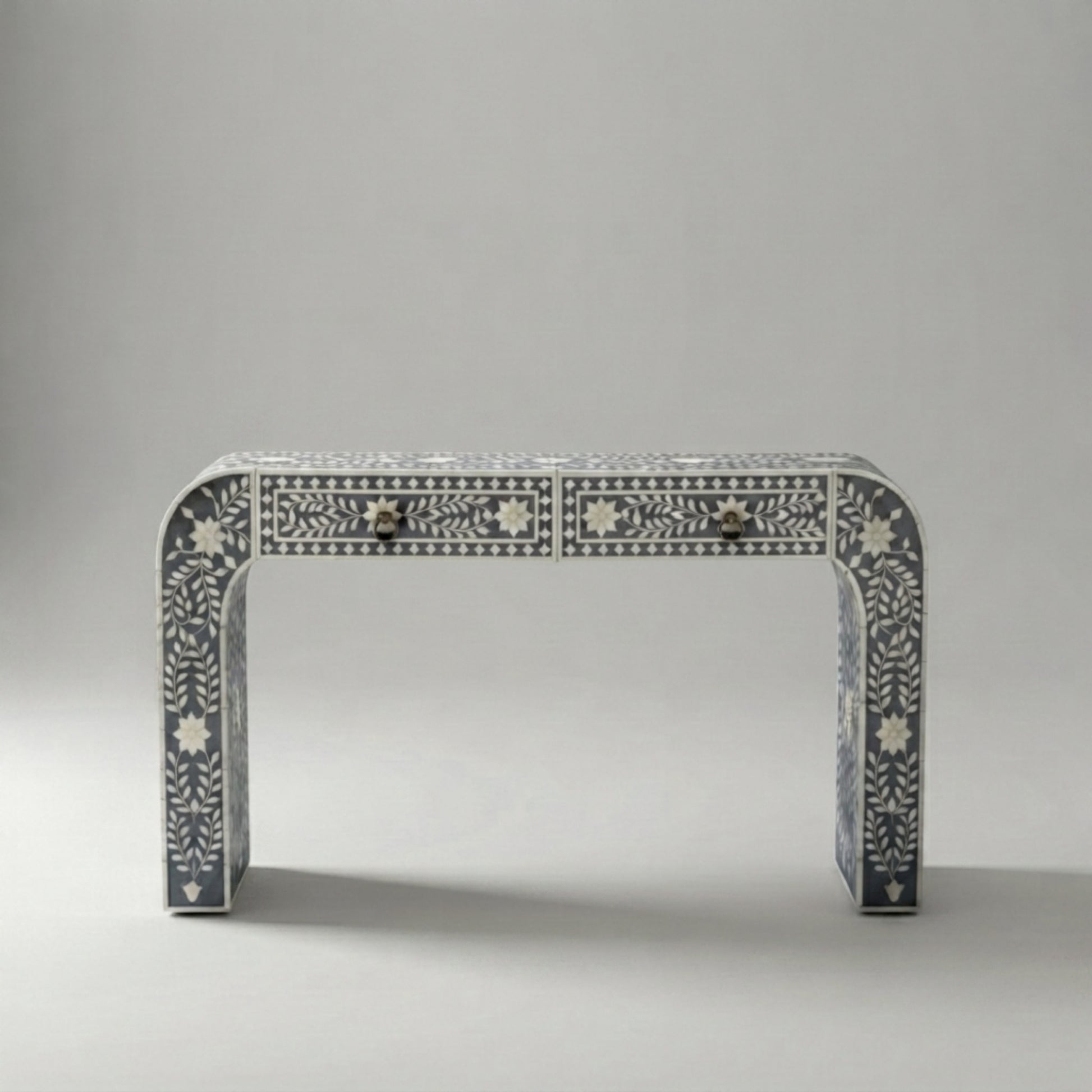 A console table with a curved top and legs, decorated with intricate white bone or mother-of-pearl inlay patterns on a dark gray base. It features two small drawers at the front with dark metal knobs, set against a neutral gray studio background.