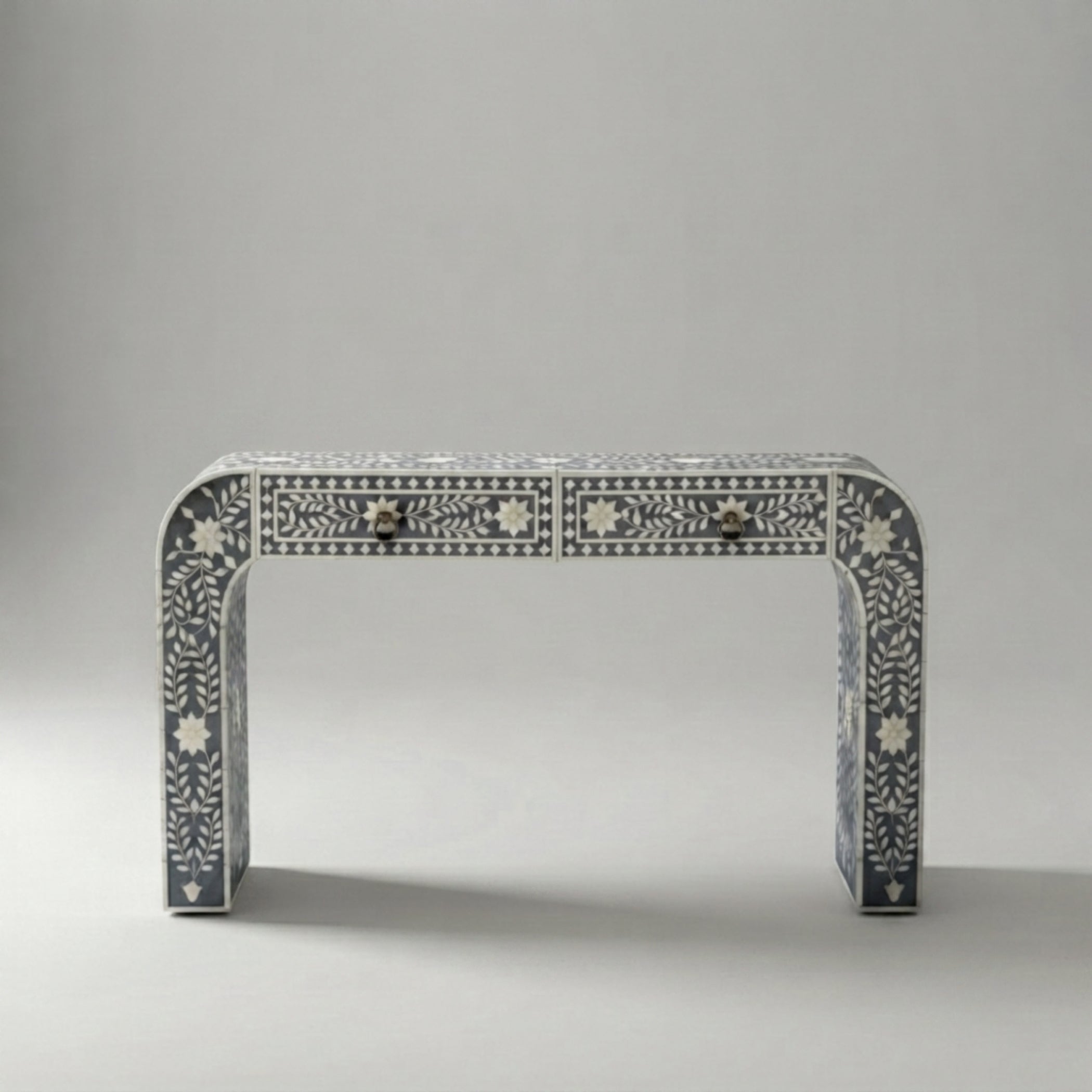 A console table with a curved top and legs, decorated with intricate white bone or mother-of-pearl inlay patterns on a dark gray base. It features two small drawers at the front with dark metal knobs, set against a neutral gray studio background.