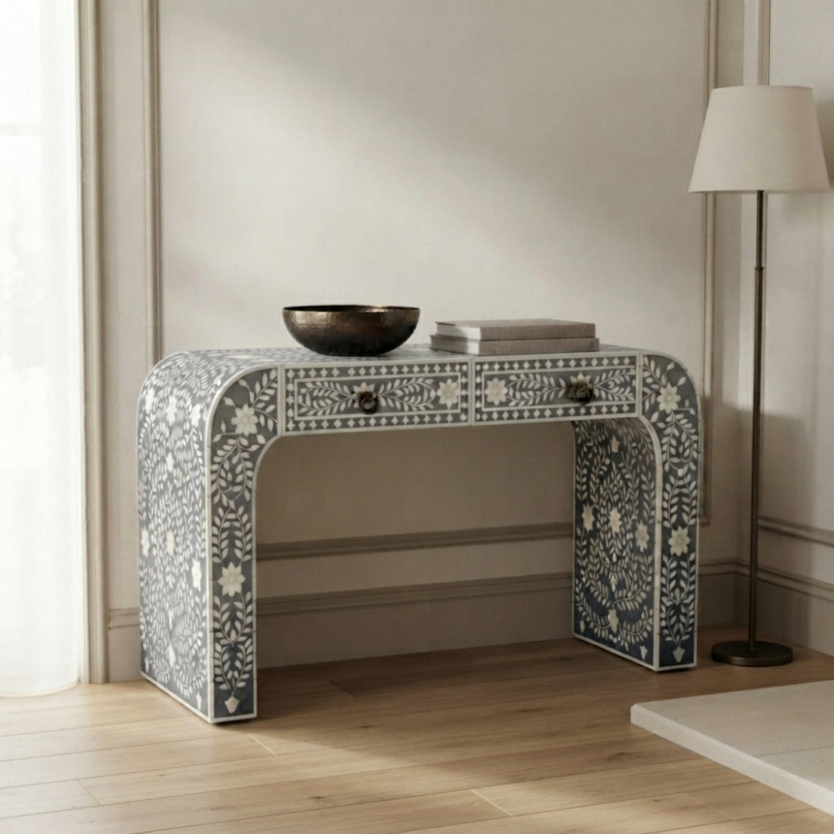 A decorative console table with intricate white floral inlay on a gray base, styled in a bright room with a floor lamp, books, and a bowl on top.