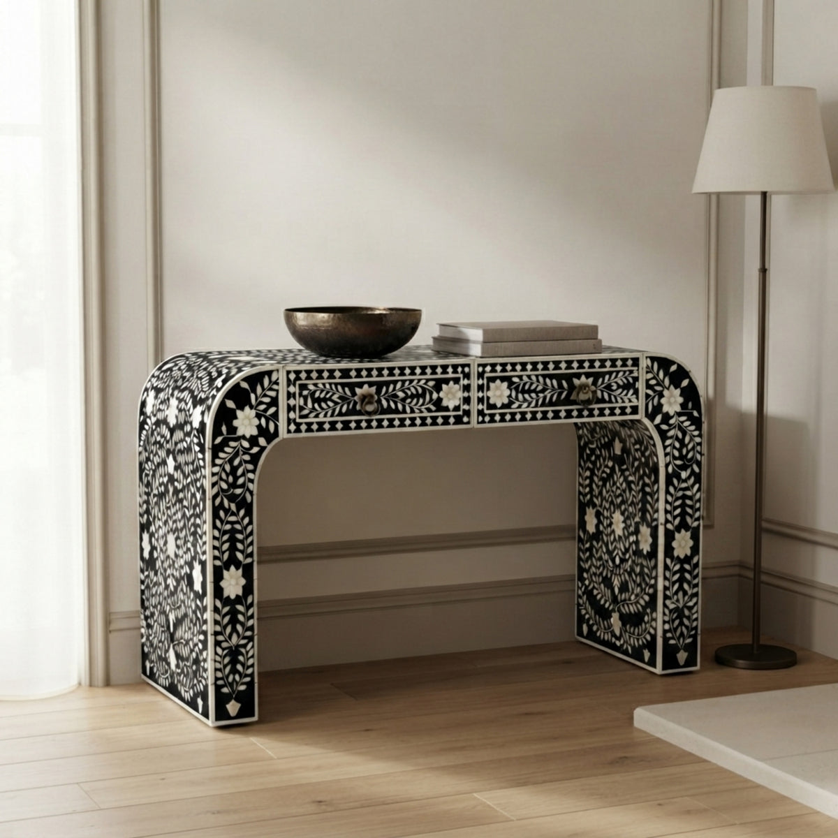 A black and white bone-inlay console table styled in a bright, modern room with light wood floors, a floor lamp, and a window curtain.