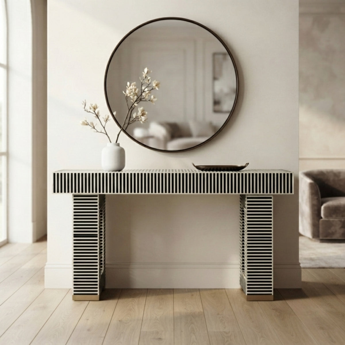 A modern console table with black and white striped bone-look legs and top, styled in a bright entryway with a round wall mirror, white vase with branches, and a small bowl.