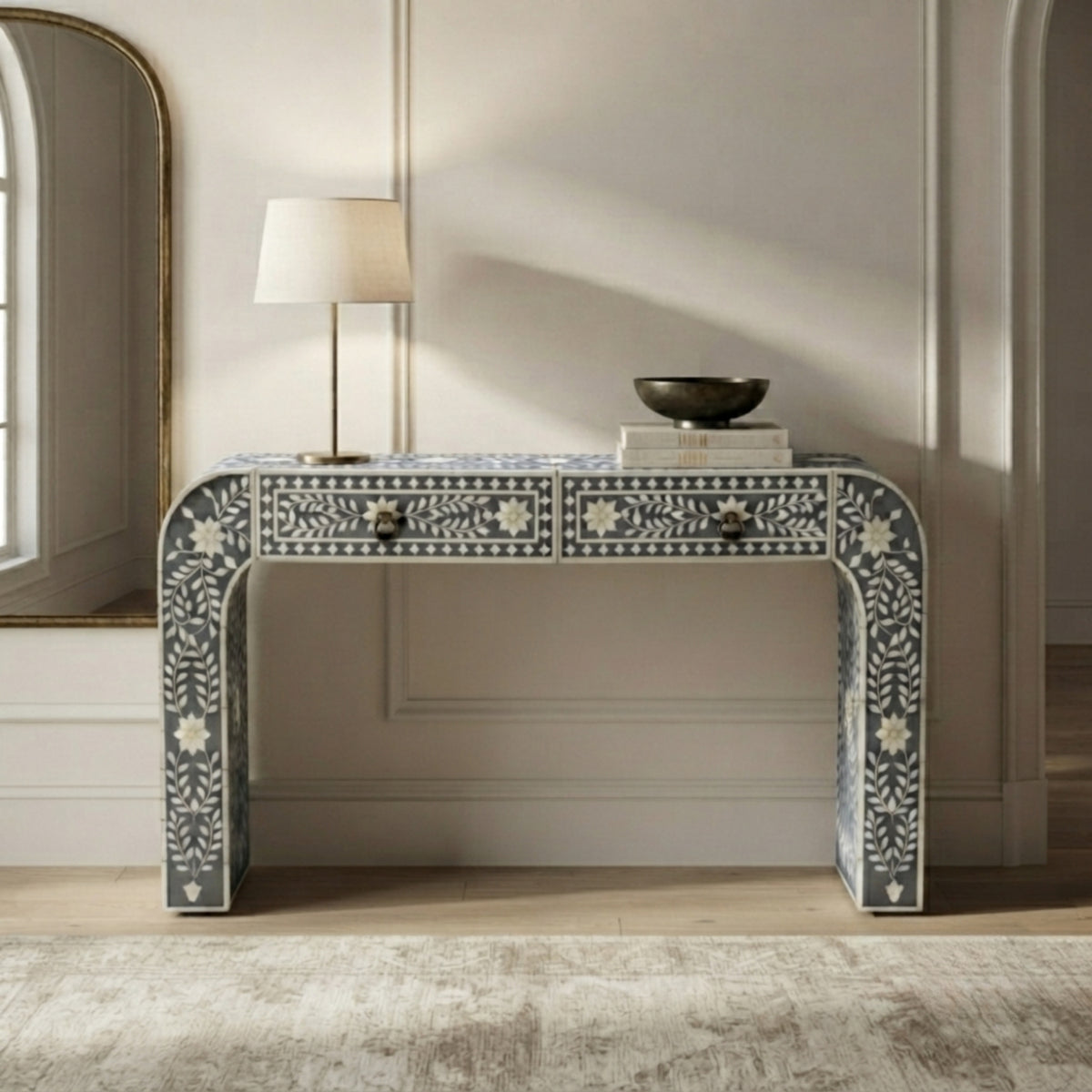 A decorative console table with two drawers, featuring intricate white floral and geometric bone-inlay patterns on a dark gray base. The table is styled in a bright, elegant room with a white table lamp, a small bowl, and books on top, set against a cream paneled wall.