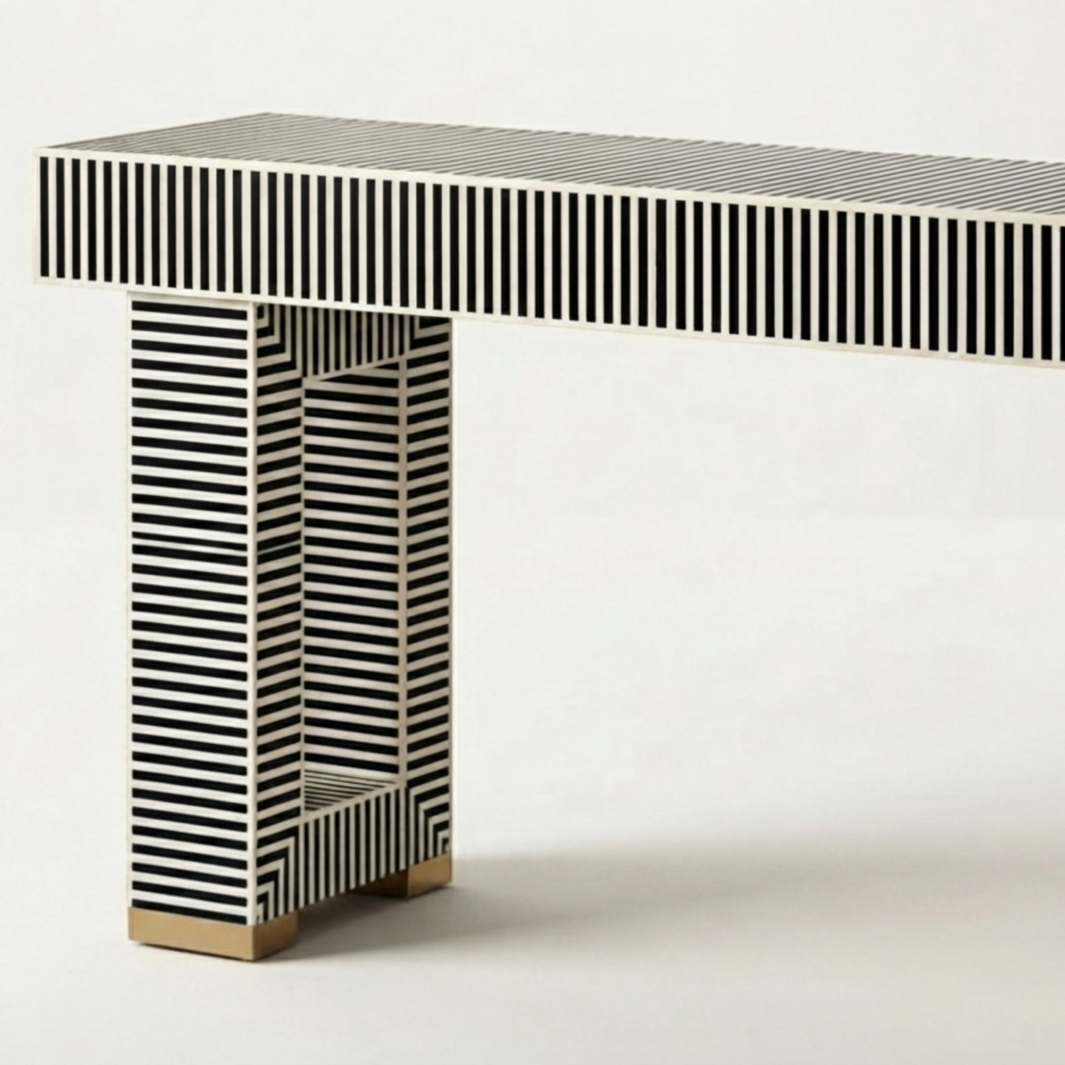 A modern console table with a rectangular top and column-style legs, both covered in black and white vertical stripes, with small gold-toned metal feet at the bottom, shown on a light gray studio background.