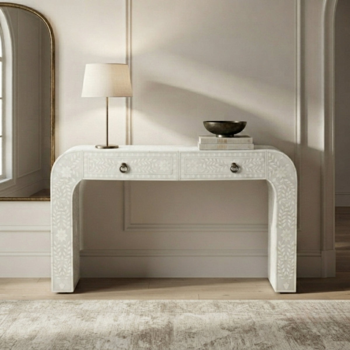 A white carved console table with two drawers and round silver knobs, styled in a bright room with a table lamp, books, and a bowl on top.