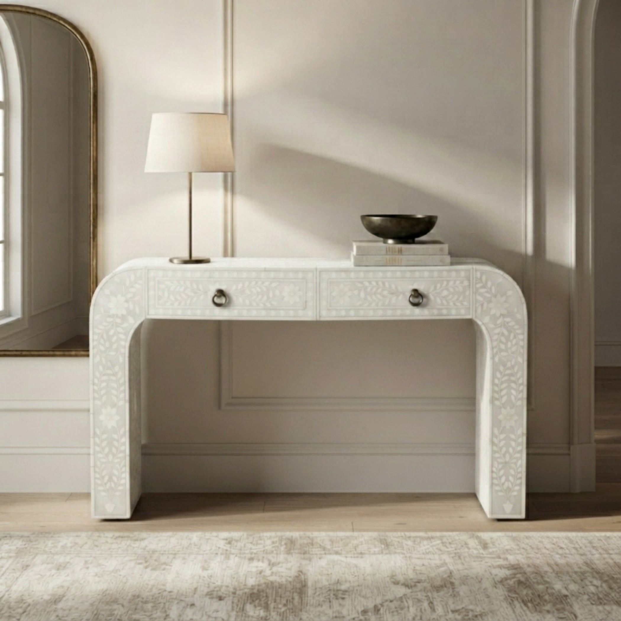 A white carved console table with two drawers and round silver knobs, styled in a bright room with a table lamp, books, and a bowl on top.