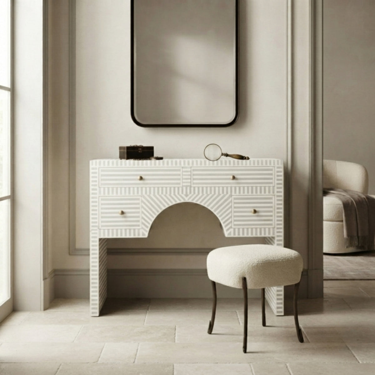 A white carved vanity table with a matching upholstered stool in a neutral-toned room, featuring a black-framed mirror and a small desk accessory on top.