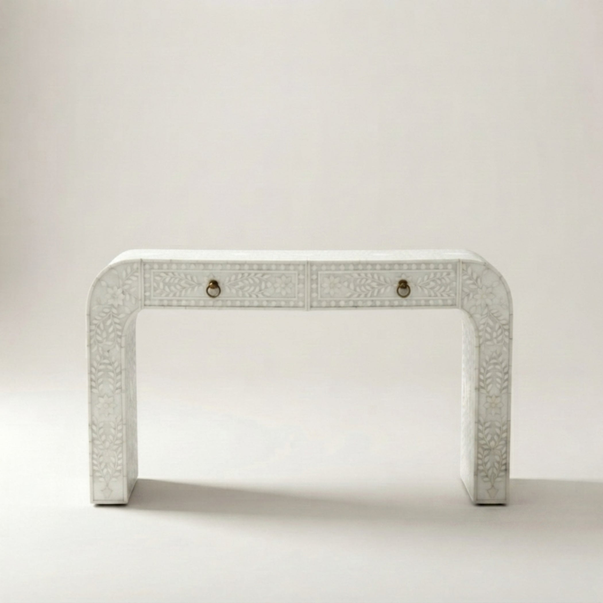 A white carved console table with two drawers and small brass pull handles, set against a neutral light gray background.