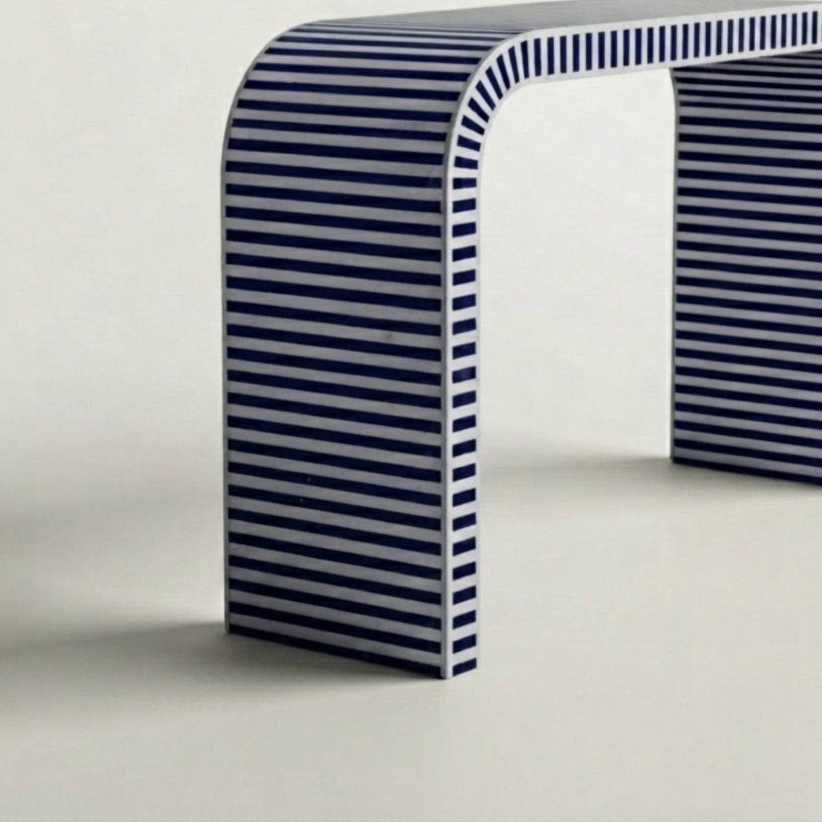 A modern curved bench with a bold navy blue and white horizontal striped pattern, shown from a low angle against a clean off-white background.
