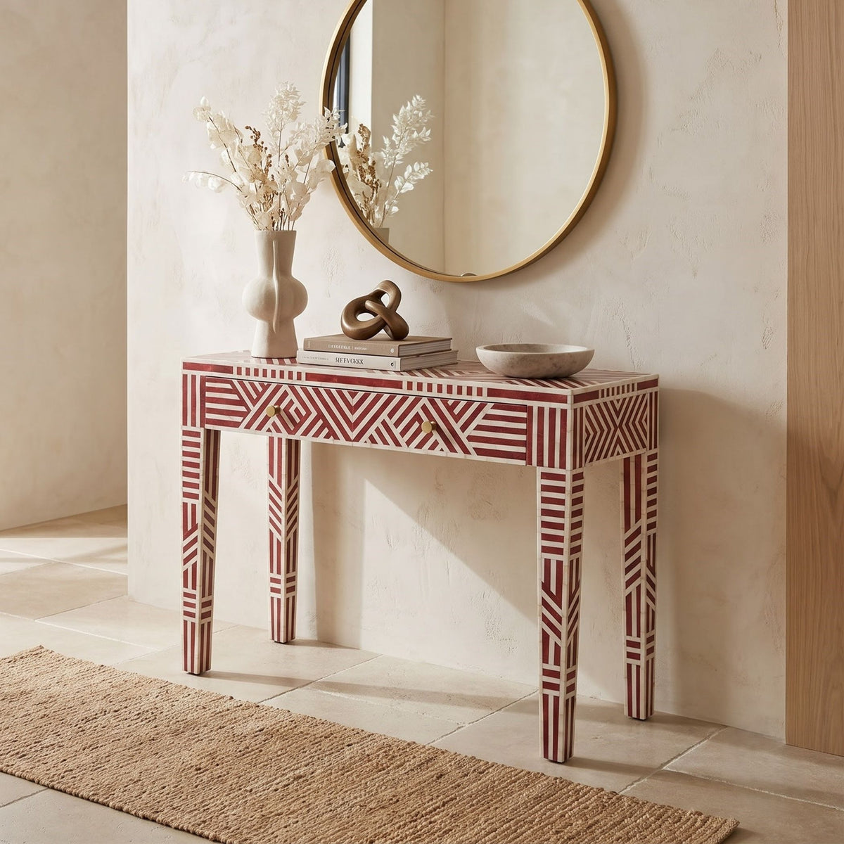 A red and white geometric patterned console table styled in a warm, neutral-toned entryway with a round gold-framed mirror, a vase of dried branches, books, and decorative objects on top.