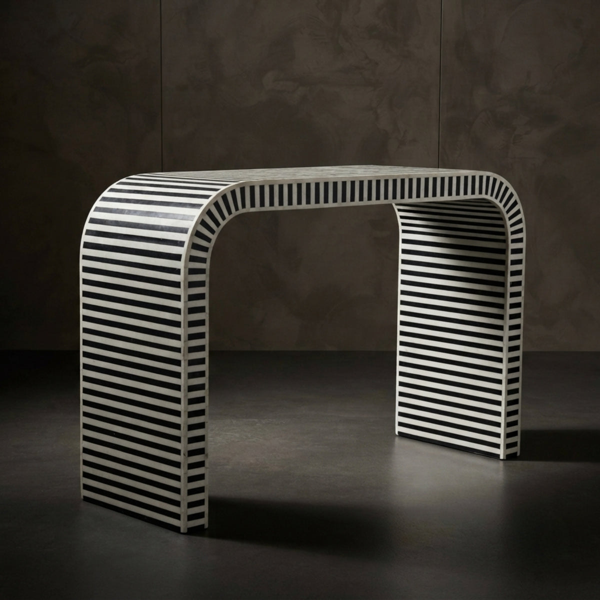 A modern console table with a curved top and legs, featuring a black and white striped bone-inlay pattern, set against a dark textured studio background.