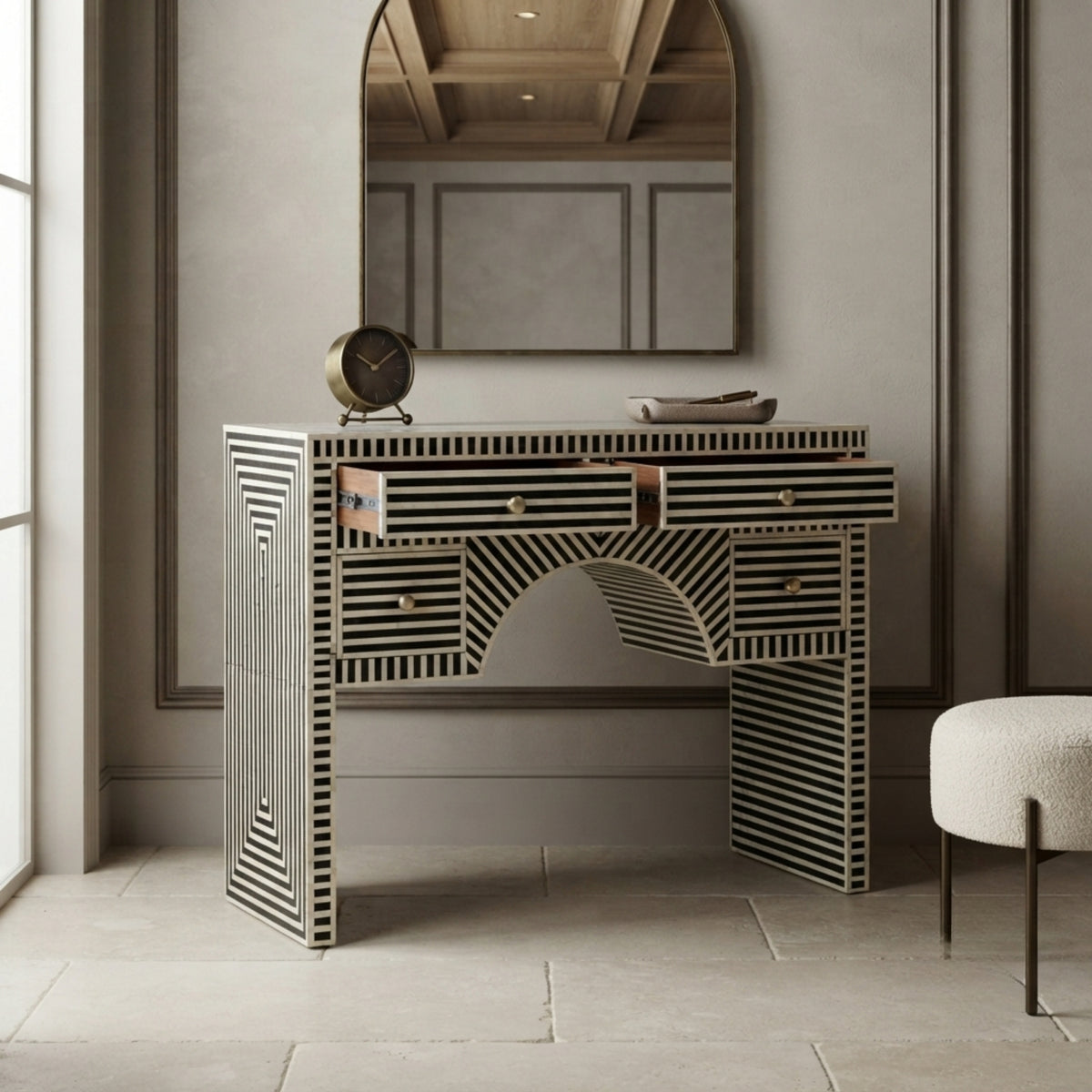 A console table with a black and white striped bone-inlay design, styled in a modern interior setting with a mirror, clock, and stool.