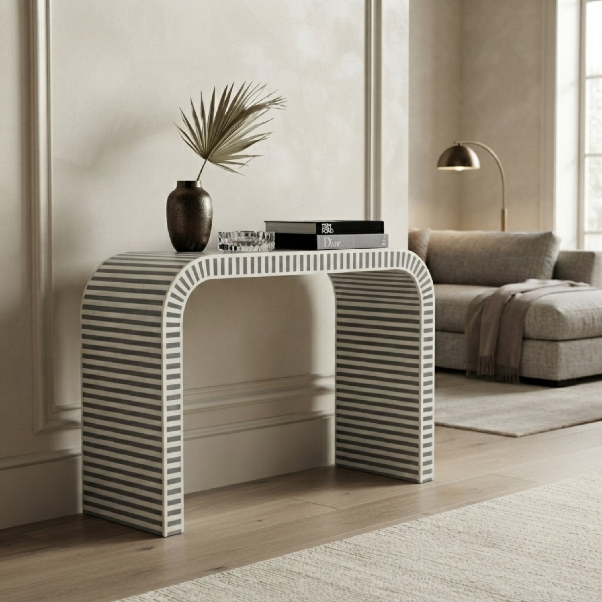 A modern console table with a curved top and striped legs in white and dark gray, styled in a neutral living room setting with a vase, books, and a sofa in the background.