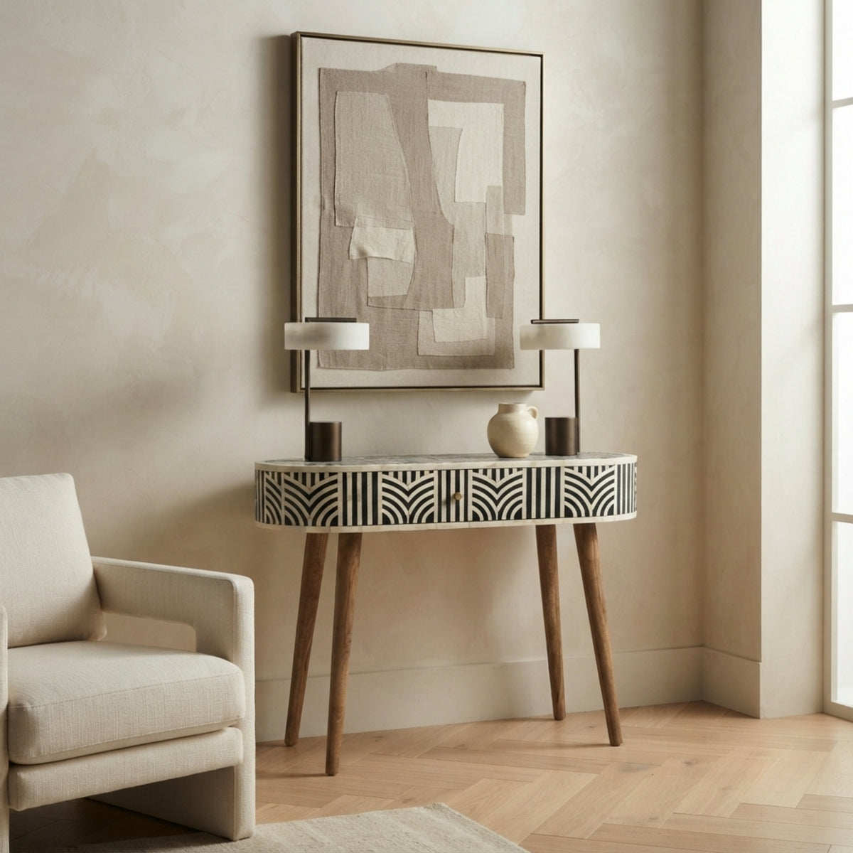 A modern console table with a black and white geometric bone-inlay pattern and tapered wooden legs, styled in a warm beige entryway with two table lamps, a ceramic vase, and a framed abstract print on the wall.