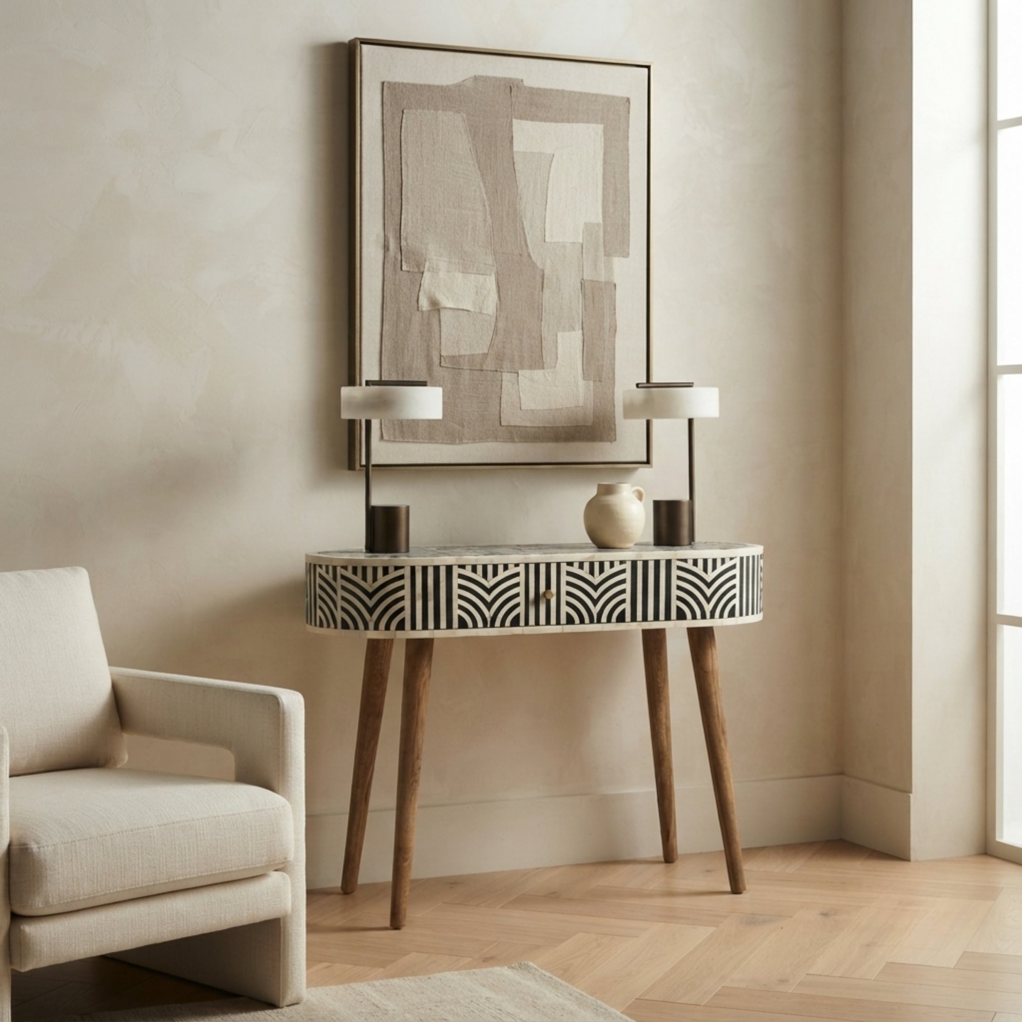 A modern console table with a black and white geometric bone-inlay pattern and tapered wooden legs, styled in a warm beige entryway with two table lamps, a ceramic vase, and a framed abstract print on the wall.