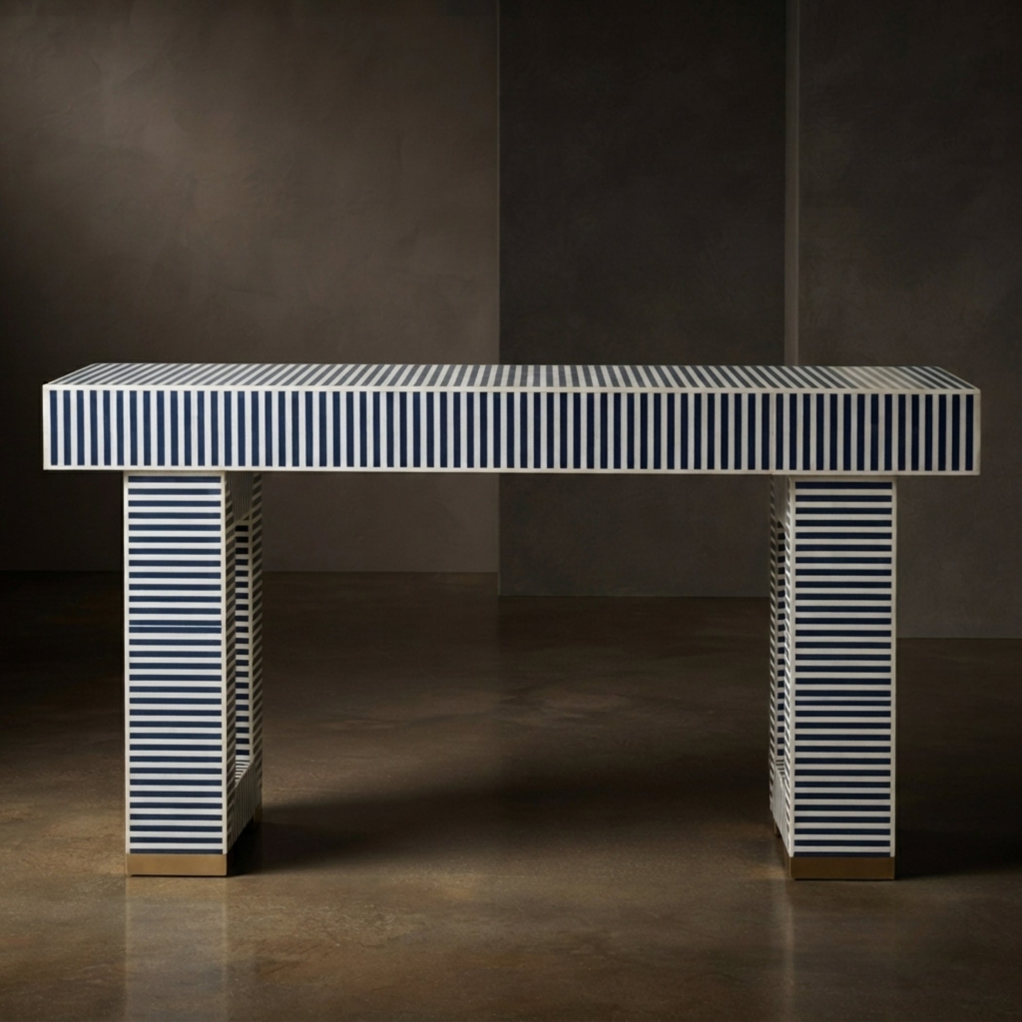 A modern rectangular console table with a white top and legs featuring vertical navy blue and white stripes, finished with small gold metal feet, set against a dark textured studio background.