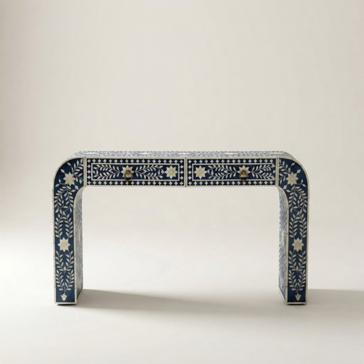 A console table with a curved top and legs, decorated with intricate white bone-inlay patterns on a dark blue base, featuring two drawers with brass handles, set against a neutral light gray background.