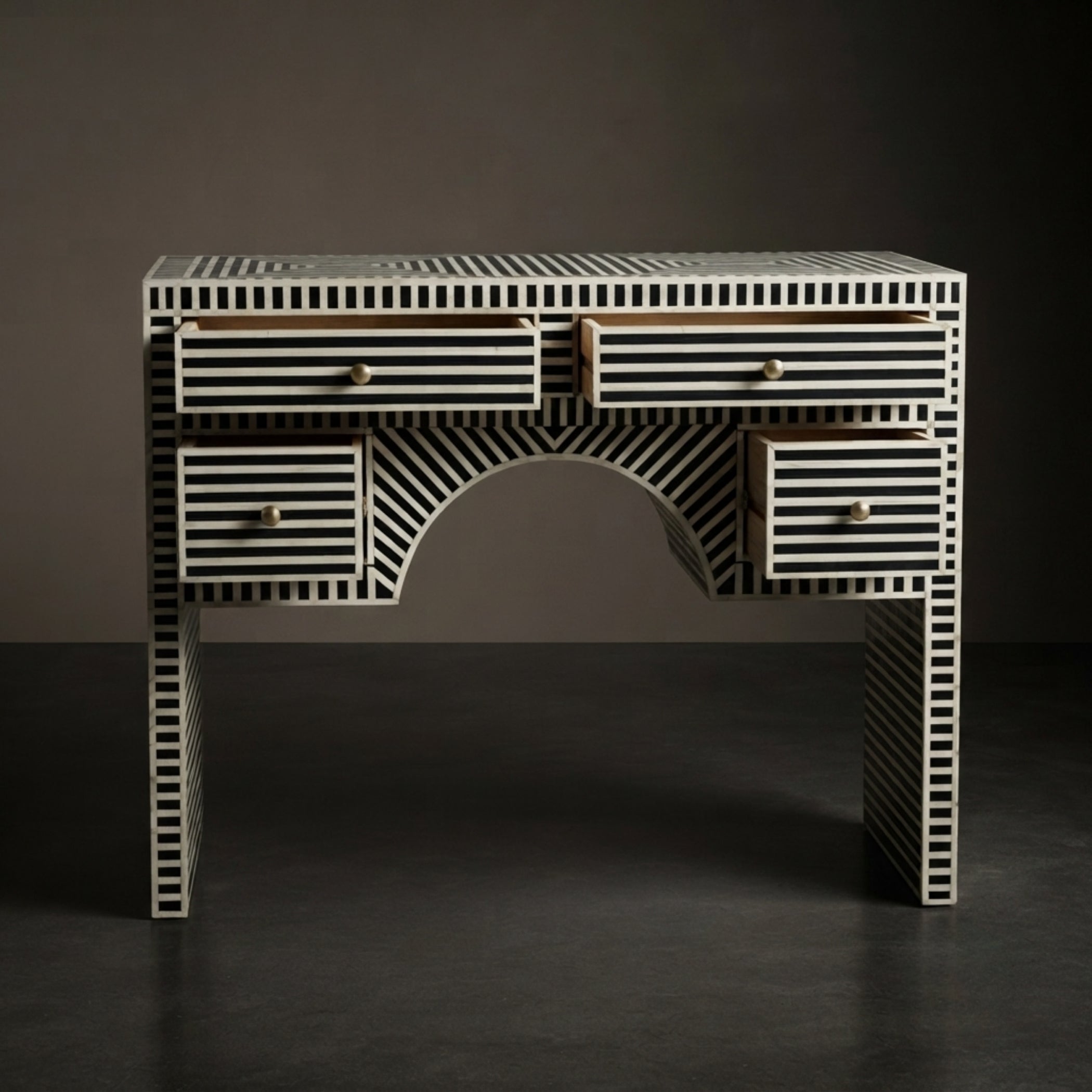 A bone-and-wood writing desk featuring a black and white striped bone inlay pattern, with a central arched opening and small brass drawer pulls, set against a dark gray background.