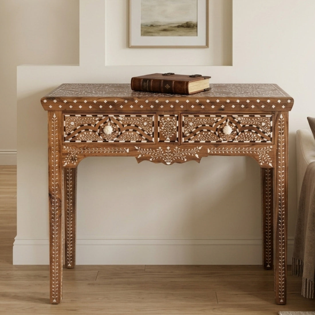 An ornate wooden console table with intricate mother-of-pearl inlay and carved details, featuring two drawers with decorative pulls, shown in a styled room setting with a book on top.