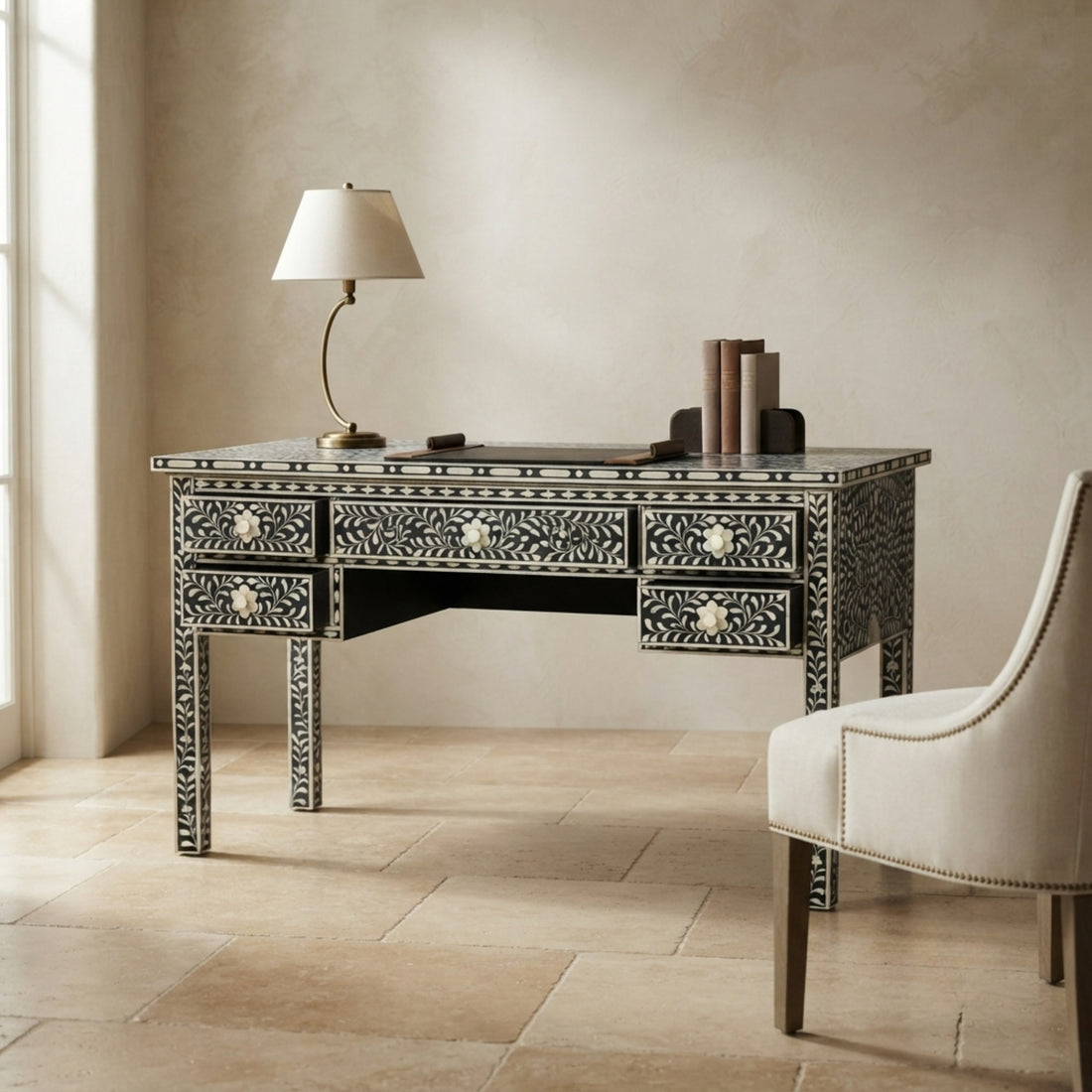 A black and white bone-inlay writing desk styled in a warm, neutral-toned room next to a cream upholstered chair, with a table lamp on top.
