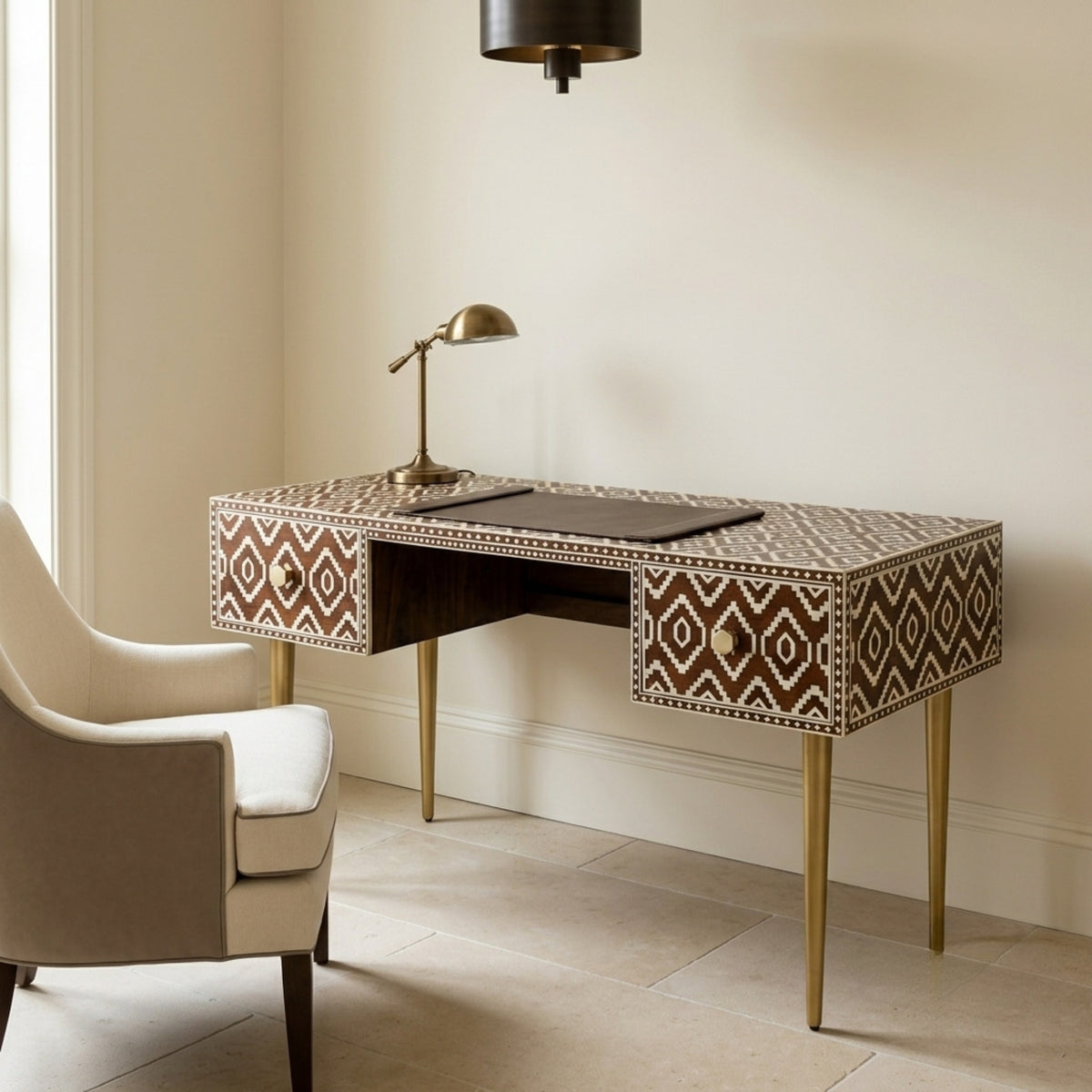 A writing desk with a brown and beige geometric bone-inlay pattern and gold legs, styled in a warm, neutral-toned room next to a beige armchair.