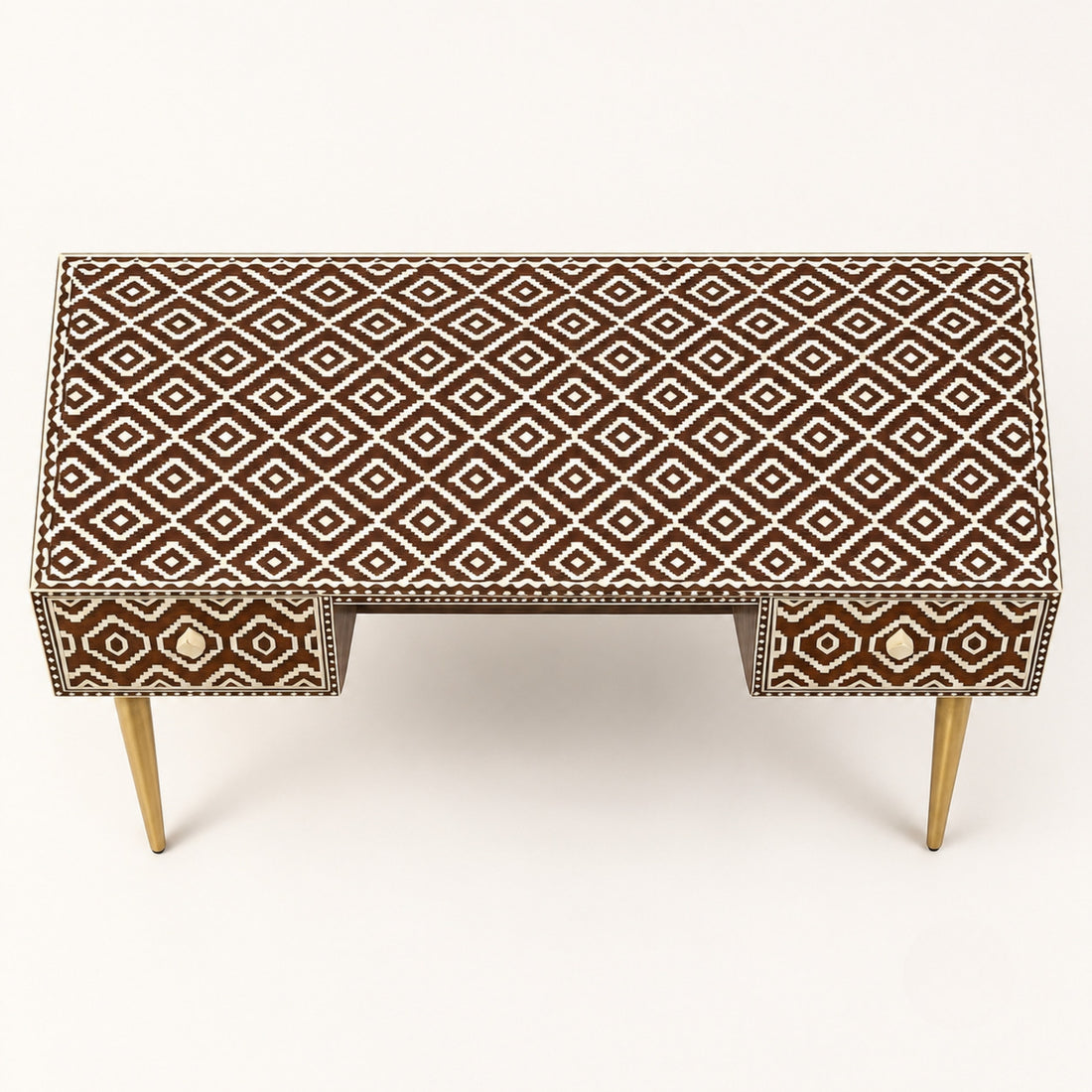 A writing desk with a brown and beige geometric bone-inlay pattern and gold legs, styled in a warm, neutral-toned room next to a beige armchair.