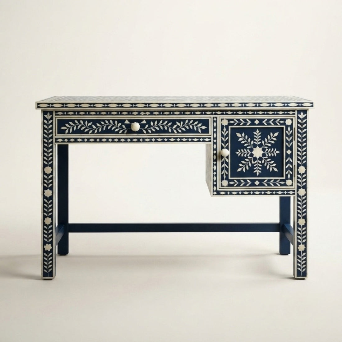 A dark blue writing desk with intricate white floral and geometric bone inlay, styled in a classic room setting with a lamp, mirror, and books.