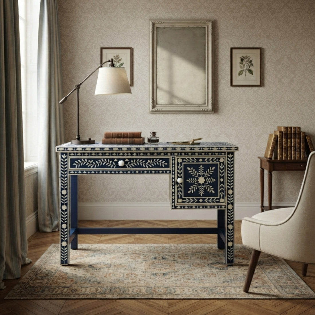 A dark blue writing desk with intricate white floral and geometric bone inlay, styled in a classic room setting with a lamp, mirror, and books.