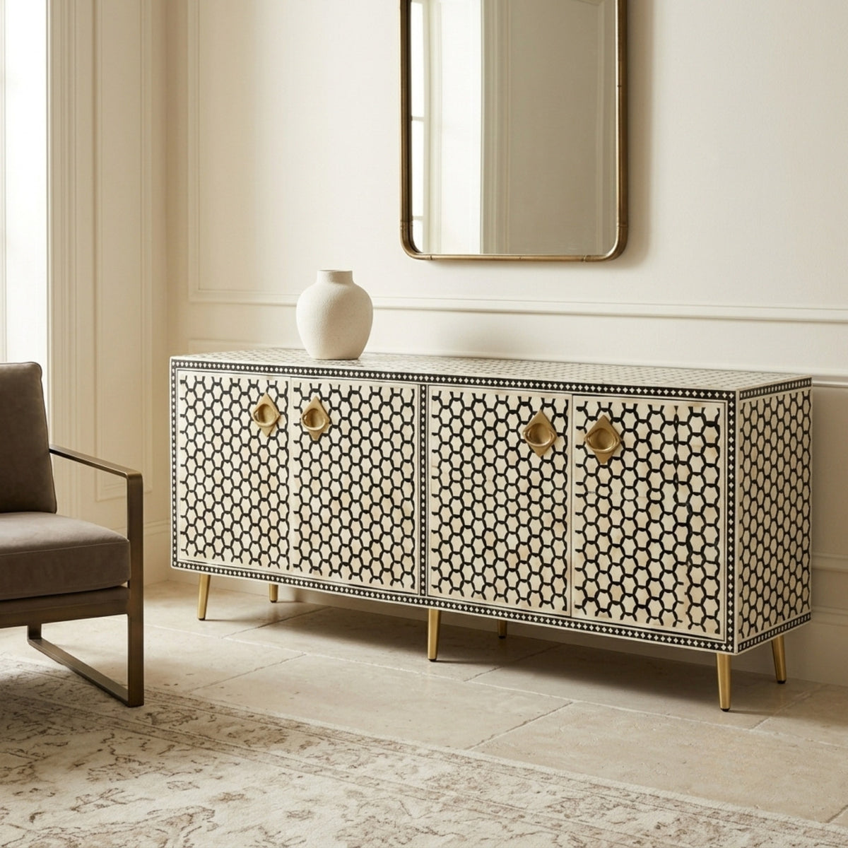 A long sideboard cabinet with an ivory and black bone-inlay pattern, gold-tone round pulls, and gold legs, styled in a bright room next to a chair with a vase on top.