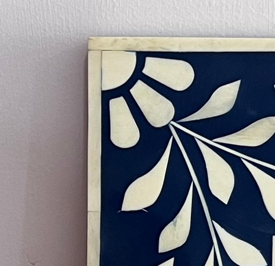 Close-up of a framed wall art piece with a navy blue background and cream floral leaf pattern, mounted on a light wall.