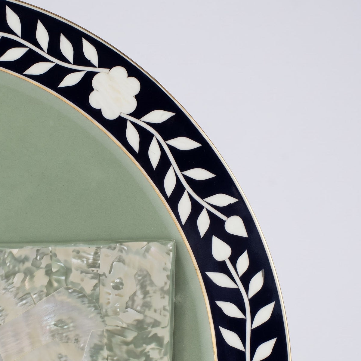 Close-up of a decorative wall mirror with a pale sage green glass center, a black border featuring white floral and leaf inlay, and a gold-toned metal frame against a light gray background.